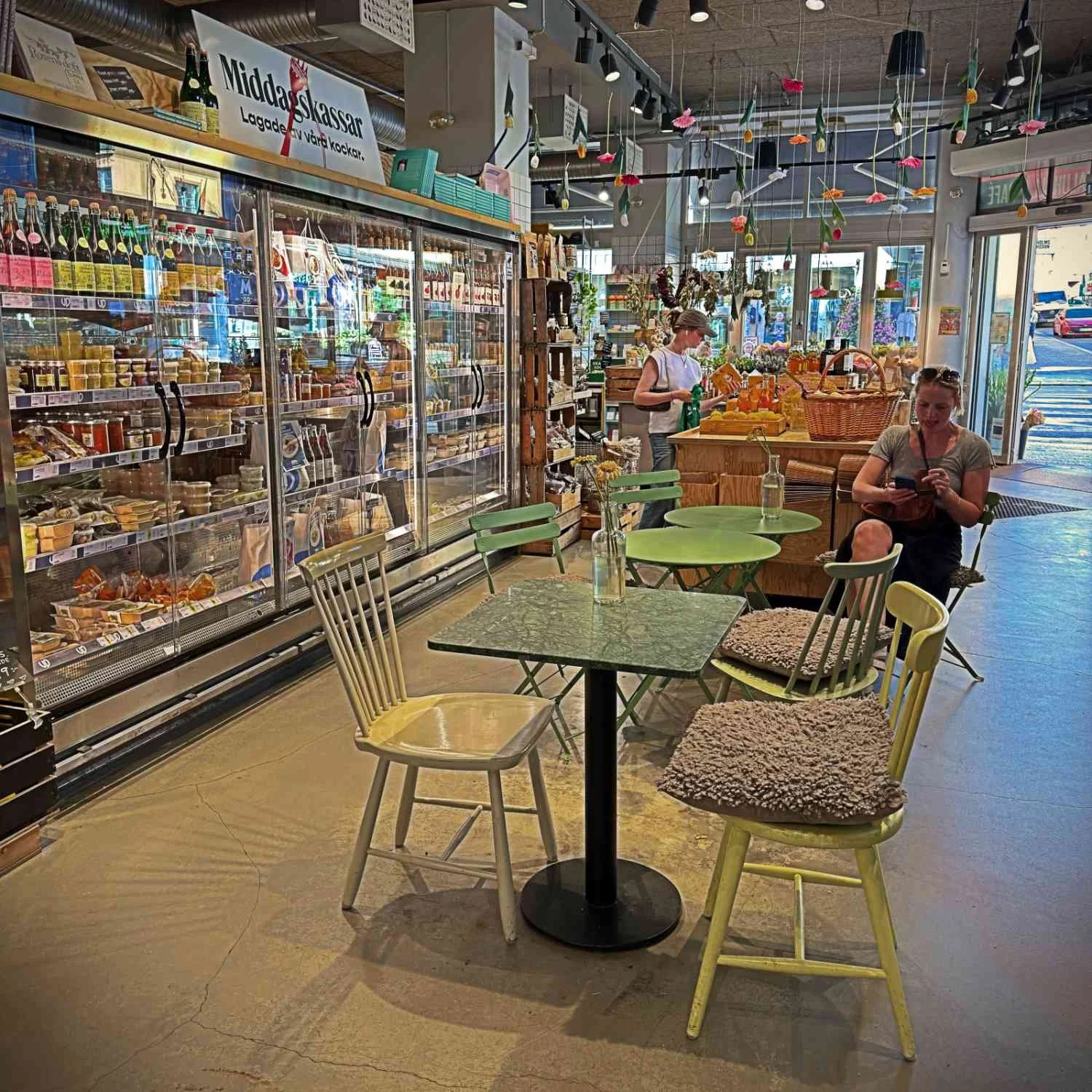 The interior seating area of Urban Deli at Nytorget featuring small bistro tables and wooden chairs. A long row of illuminated refrigerated display cases with deli products lines the left side.