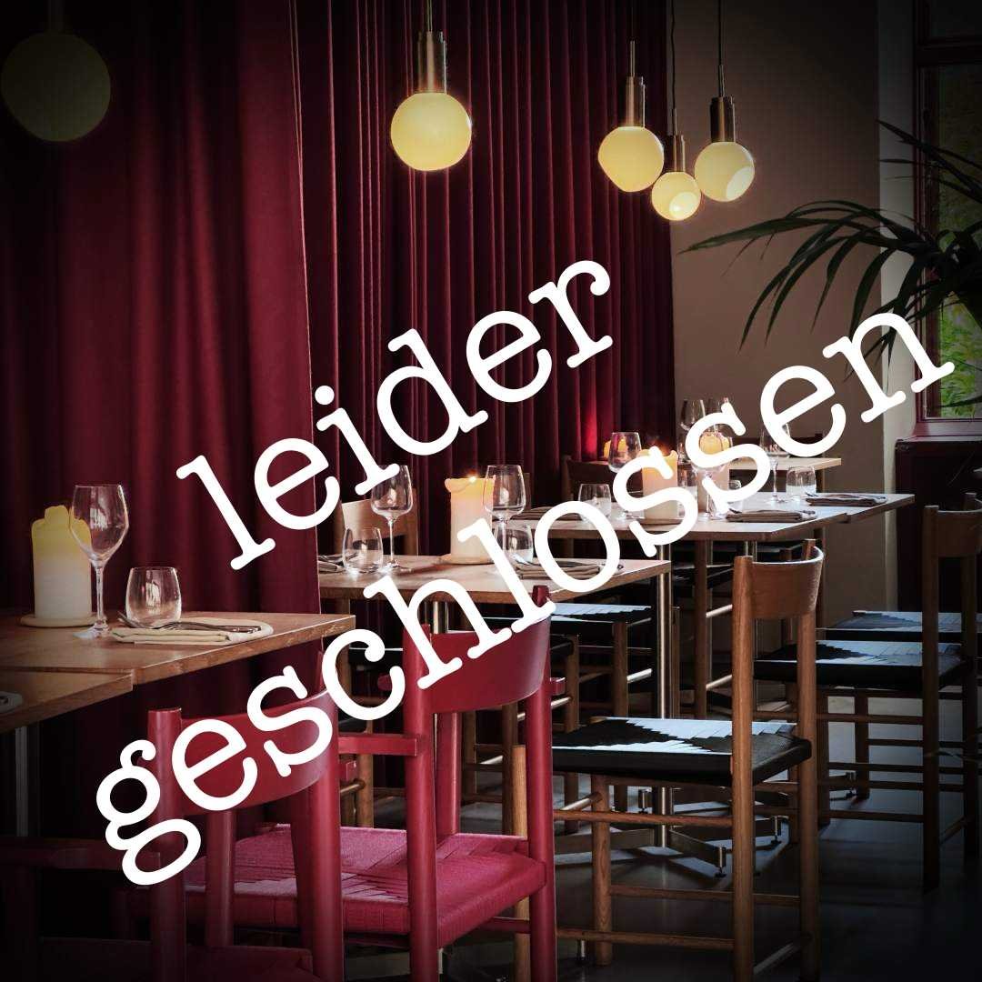 Innenaufnahme des Iluka Restaurants in Berlin mit gedeckten Holztischen, roten und schwarzen Stühlen, Kerzen und Gläsern. Im Hintergrund hängen runde Leuchten vor weinrotem Vorhang.