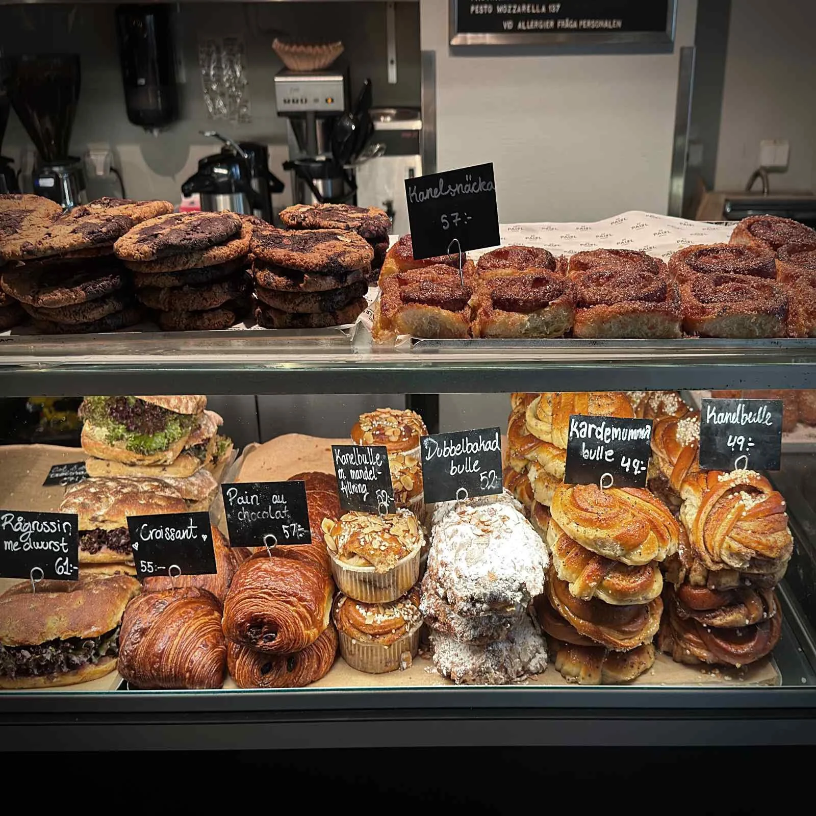 A glass display case filled with traditional Swedish baked goods, including cinnamon buns (Kanelbulle), cardamom pastries, and powdered sugar treats. Small chalkboard tags indicate the prices.