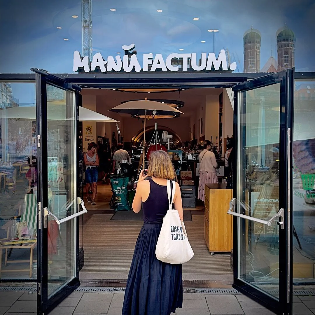 Der Eingangsbereich des Manufactum Cafés in München mit einer gläsernen Eingangstür, einem dunklen Metallrahmen und einem weißen Schriftzug auf der Glasscheibe.