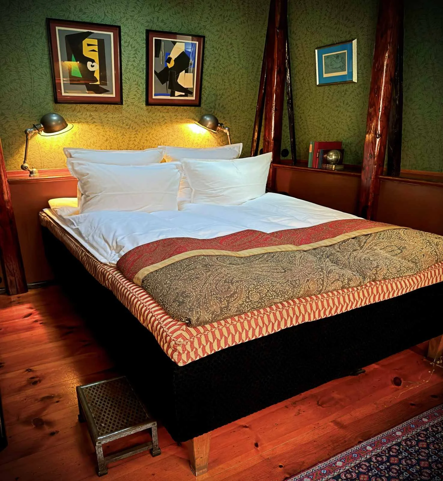 Ein Doppelbett mit weißer Bettwäsche und einer gemusterten Überdecke auf Holzdielen. Zwei eingeschaltete Leselampen flankieren das Kopfteil vor einer grünen Mustertapete mit gerahmten Bildern.