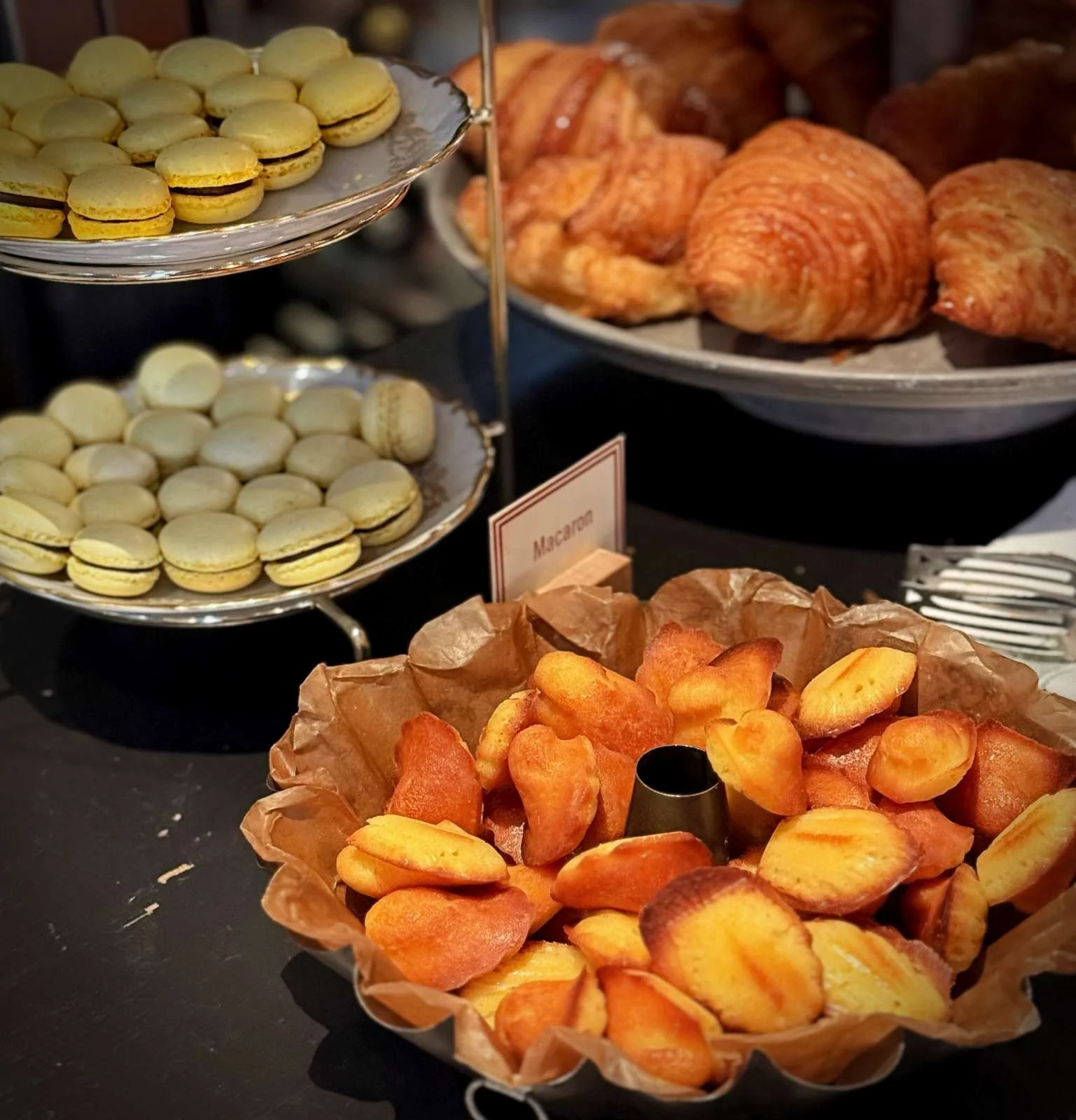 Eine Backform mit Madeleines auf Backpapier im Vordergrund. Dahinter befinden sich zwei Etagere-Teller mit gelben Macarons sowie Croissants auf einer Platte im Hintergrund. Englische Bildbeschreibung: A baking