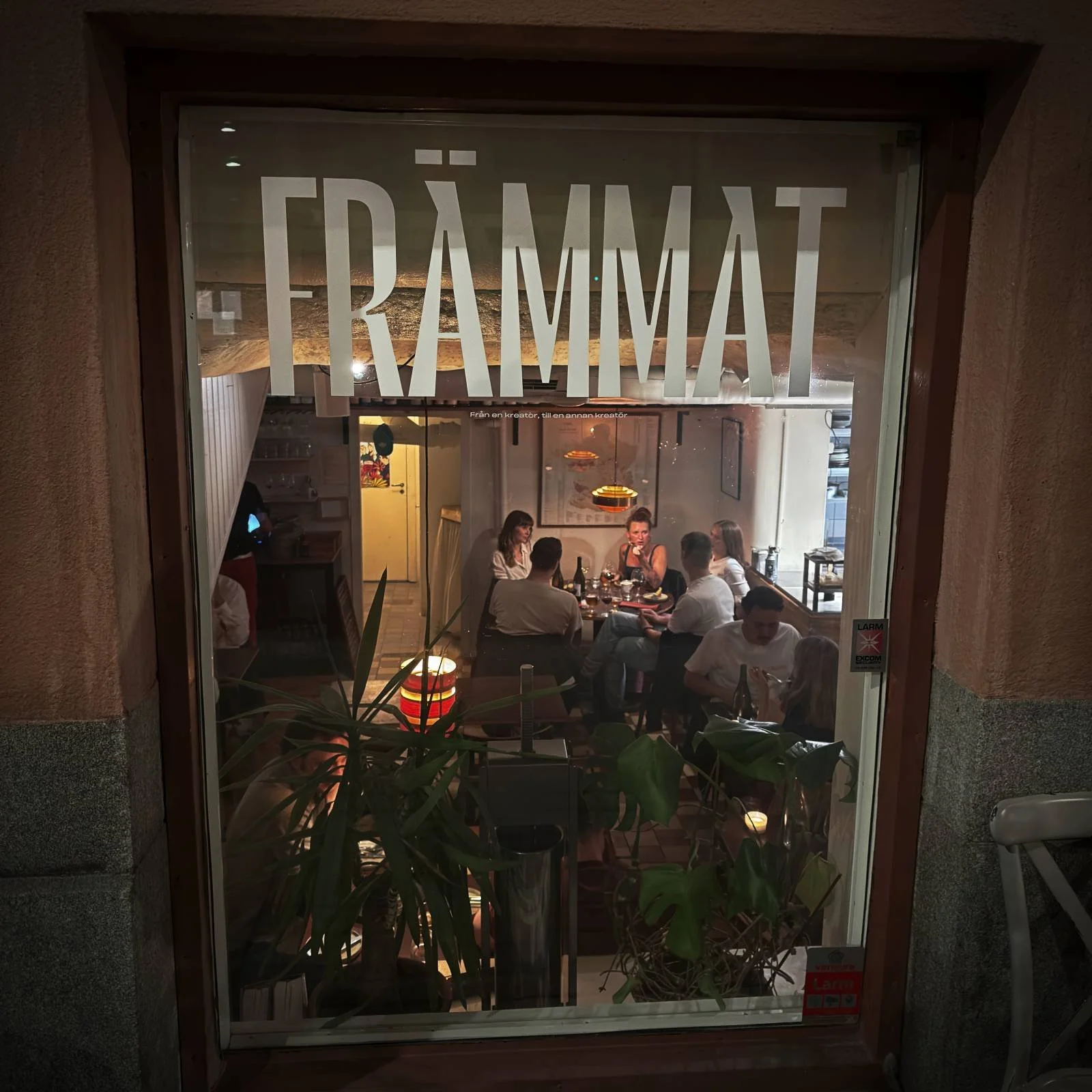 Der Blick durch das große Schaufenster des Restaurants Främmat in Stockholm zeigt Gäste, die in dem gemütlich und warm beleuchteten Innenraum an einem zentralen Tisch gemeinsam speisen.