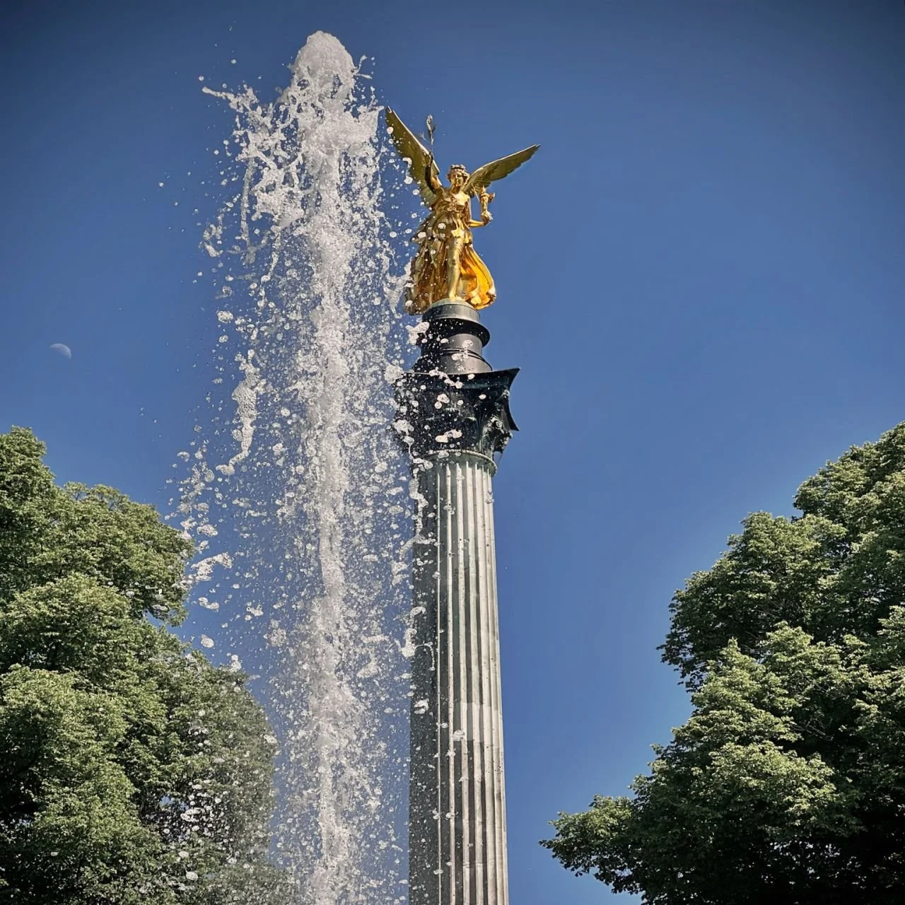 Der Friedensengel in München mit vergoldeter Statue auf hoher Säule, daneben eine Fontäne mit aufsteigendem Wasser, eingerahmt von Bäumen vor blauem Himmel.