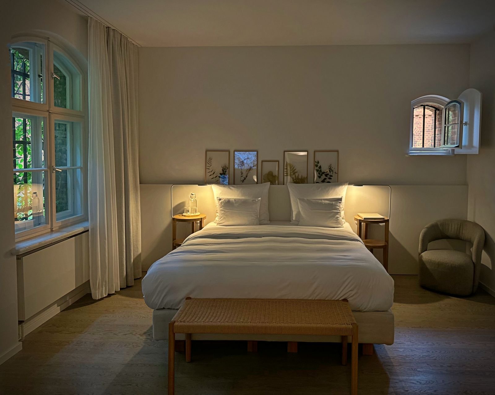 Ein Doppelbett mit weißer Bettwäsche im Hotel Wilmina in Berlin. Das schlichte Zimmer verfügt über zwei Nachttischlampen, botanische Wandbilder und ein Fenster mit Blick in den grünen Garten.
