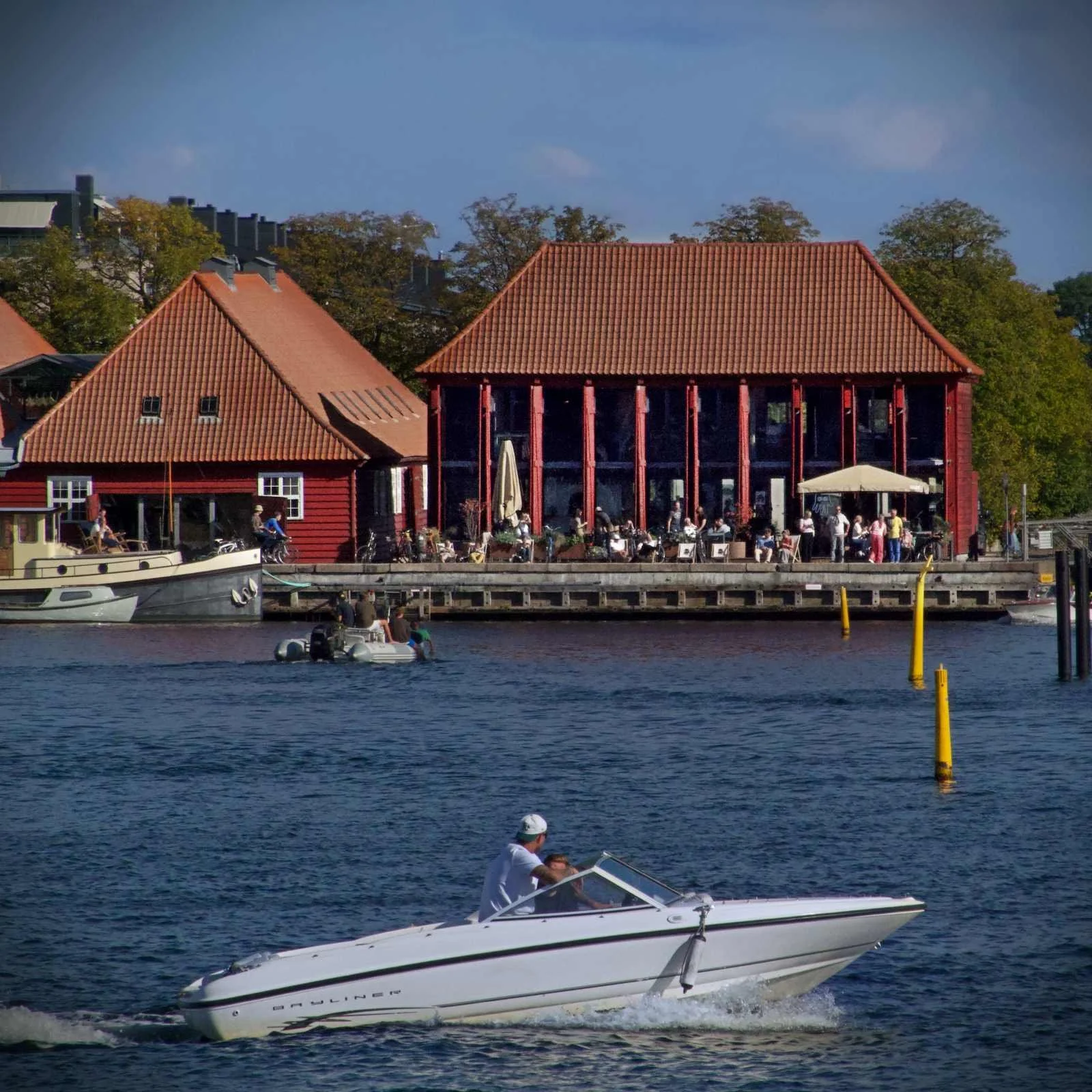 Ein weißes Motorboot fährt auf dem Wasser vor dem 25hours Hotel Paper Island in Kopenhagen. Im Hintergrund sind rote Gebäude am Hafenbecken und die Terrasse der Hart Bakery zu sehen.