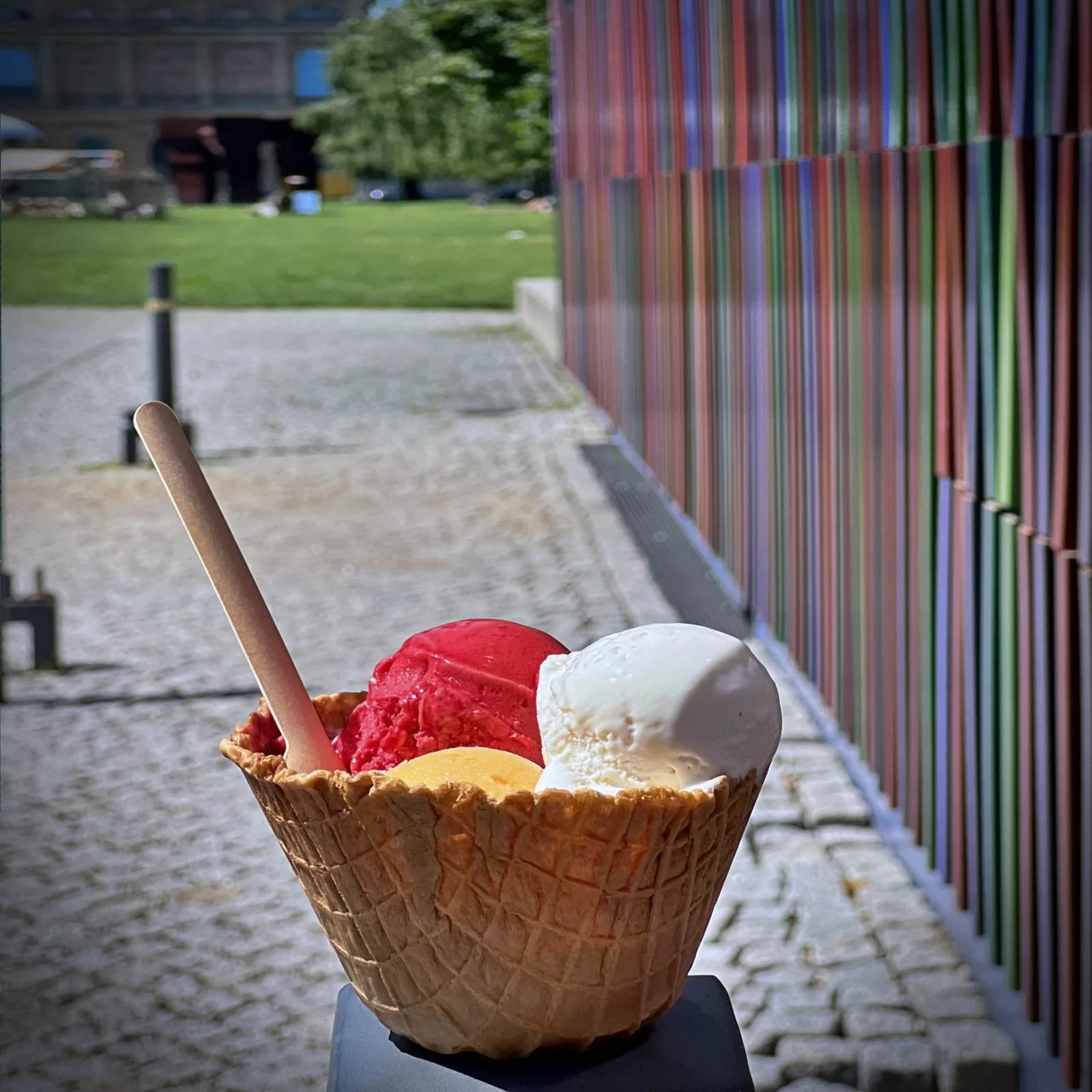 Drei Kugeln Eis in einer Waffel von Ballabeni vor der bunten Keramikfassade des Museums Brandhorst in München. Im Hintergrund ist die sonnige Wiese vor den Pinakotheken zu sehen.