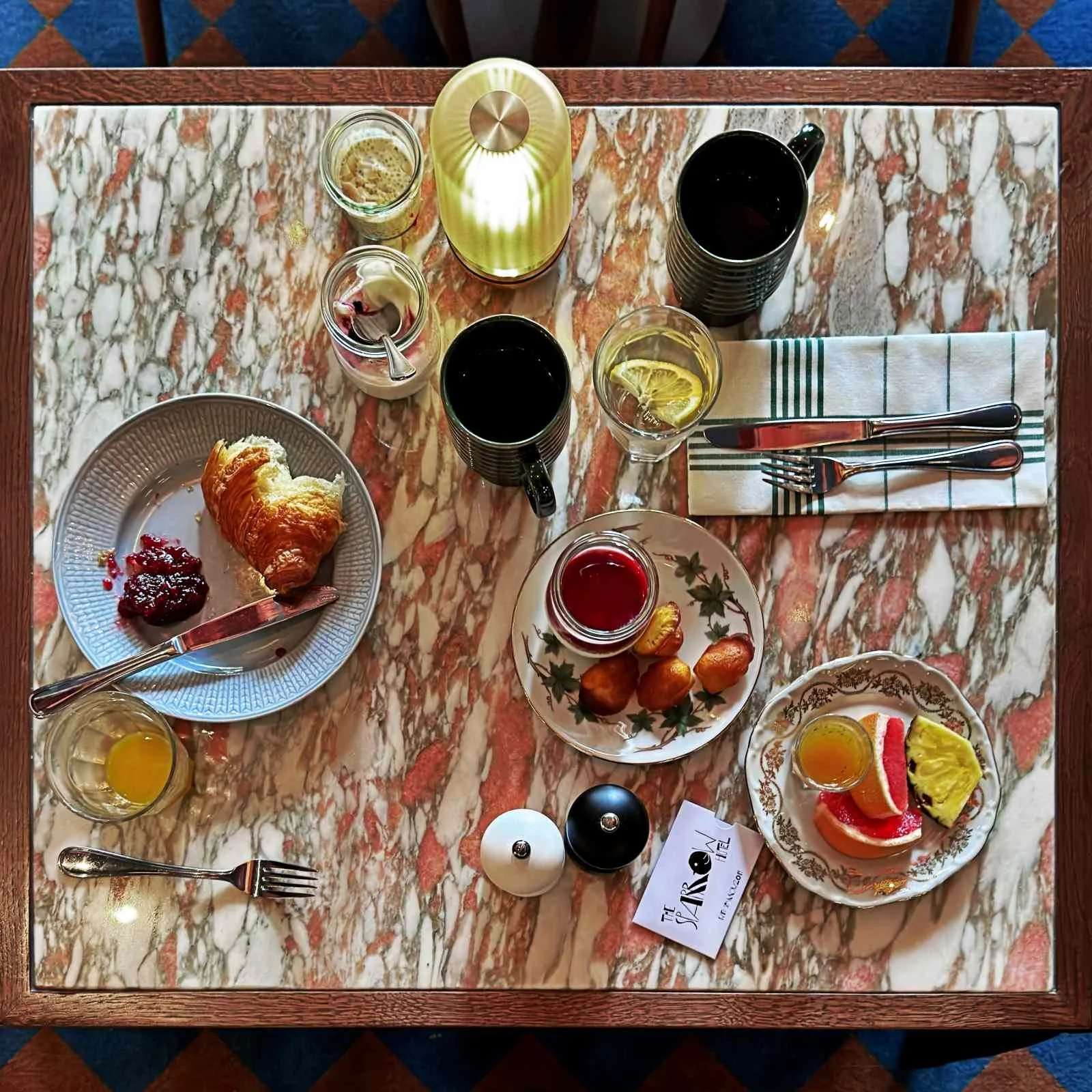 Eine Draufsicht auf einen Marmortisch mit Kaffeebechern, Joghurtgläsern, Saft, einem angeschnittenen Croissant, Madeleines und Zitrusfrüchten. Eine kabellose Tischlampe beleuchtet die Mitte.