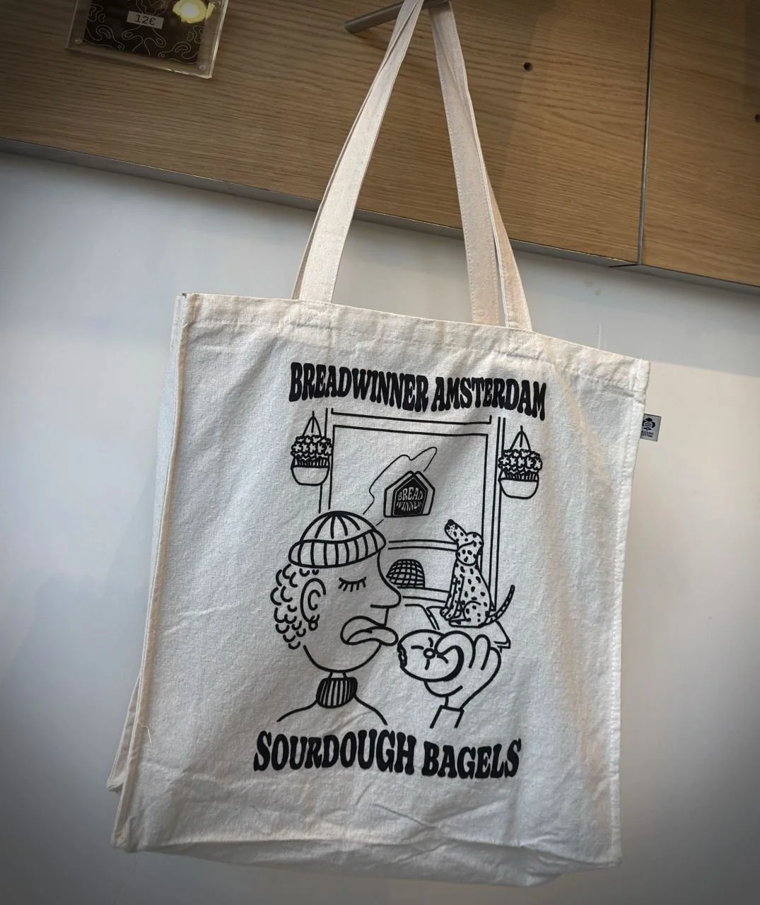 Canvas tote bag by Breadwinner Amsterdam with black illustration and “Sourdough Bagels” lettering. Cotton bag hanging on a wooden rail inside the café.