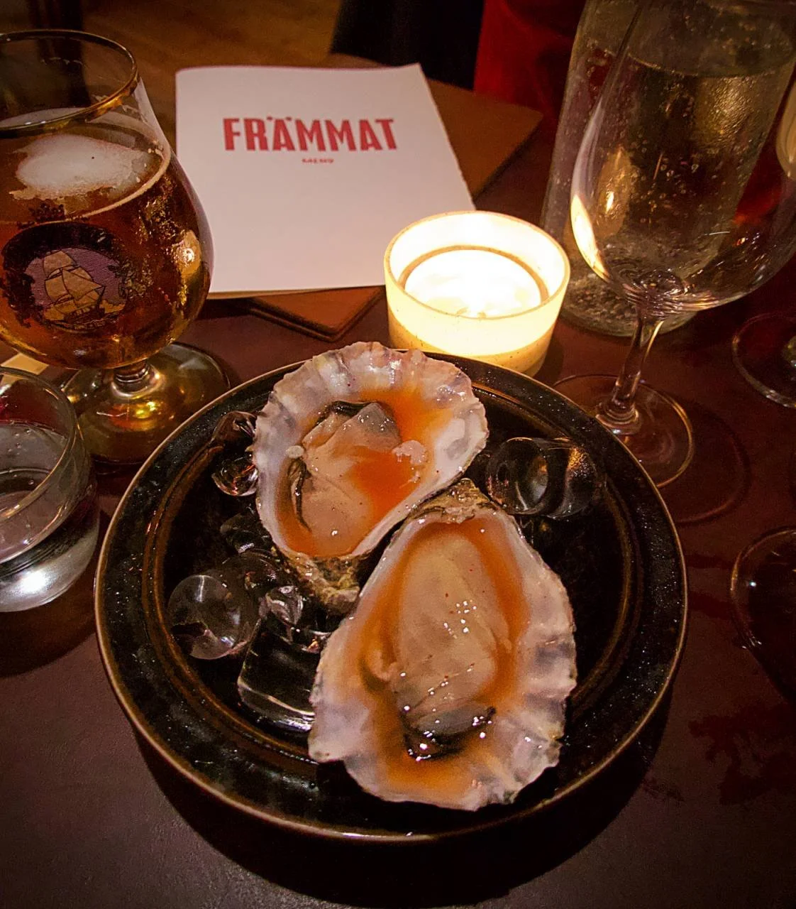 Nahaufnahme von zwei Austern auf Eis, serviert mit einer Vinaigrette im Restaurant Främmat in Stockholm, neben einem Glas Bier und Kerzenschein.
