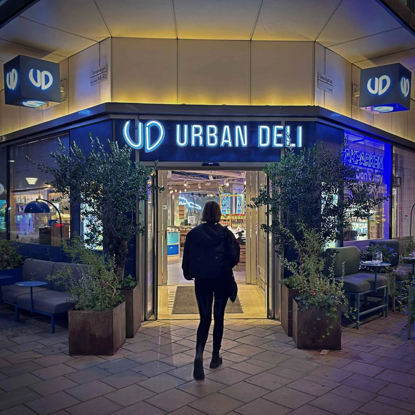 Exterior view of the Urban Deli store on Sveavägen at night, featuring a bright blue neon sign above the entrance. A person dressed in dark clothing walks through the open sliding door into the shop.