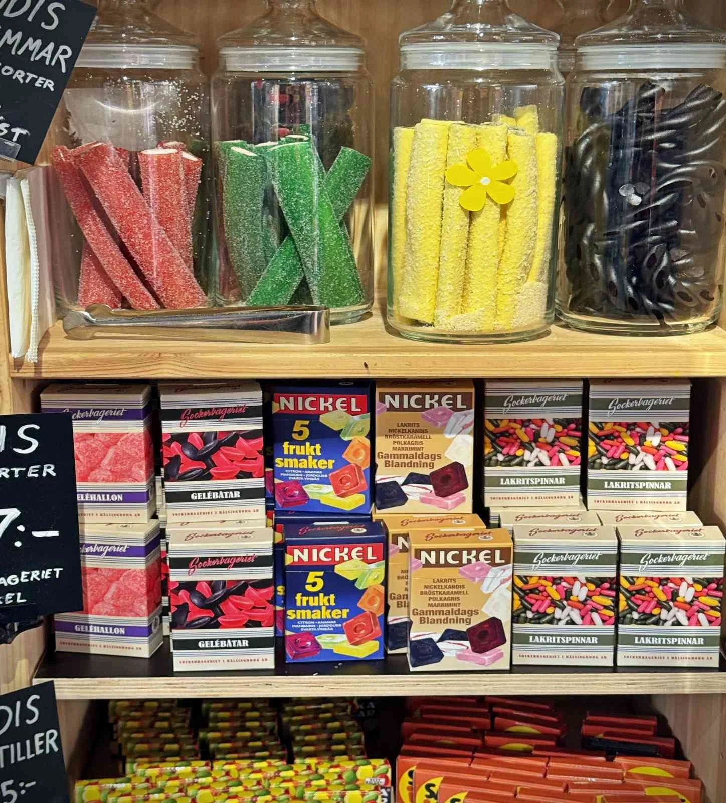 A wooden shelf inside Urban Deli filled with traditional Swedish sweets. Large glass jars with fruit and liquorice sticks sit on the top shelf, above colorful candy boxes from brands like Sockerbageriet and Nickel.
