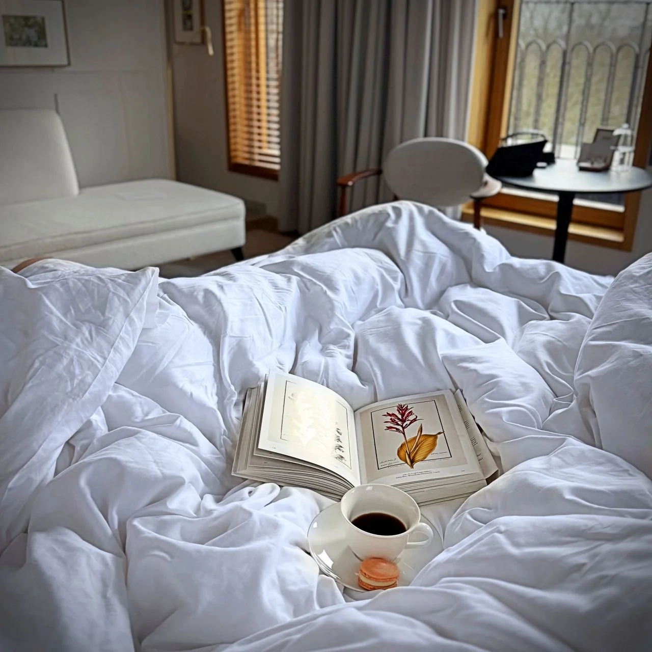 Gemütliches Hotelbett mit weißer Bettwäsche, einem aufgeschlagenen Buch, einer Tasse Kaffee und einem Macaron im Pillows Hotel Amsterdam.