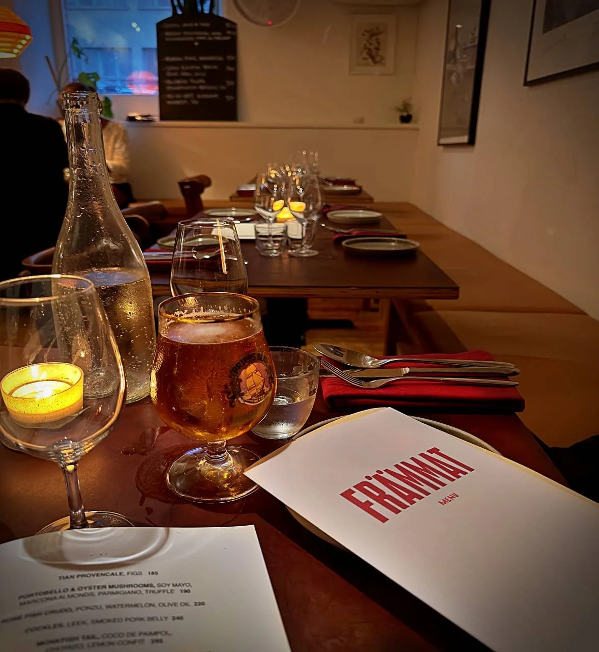 Eine  Aufnahme eines gedeckten Tisches im Restaurant Främmat in Stockholm, mit Speisekarten, einem Glas Pripps Blå Bier, Wasser und Kerzenschein im Hintergrund.