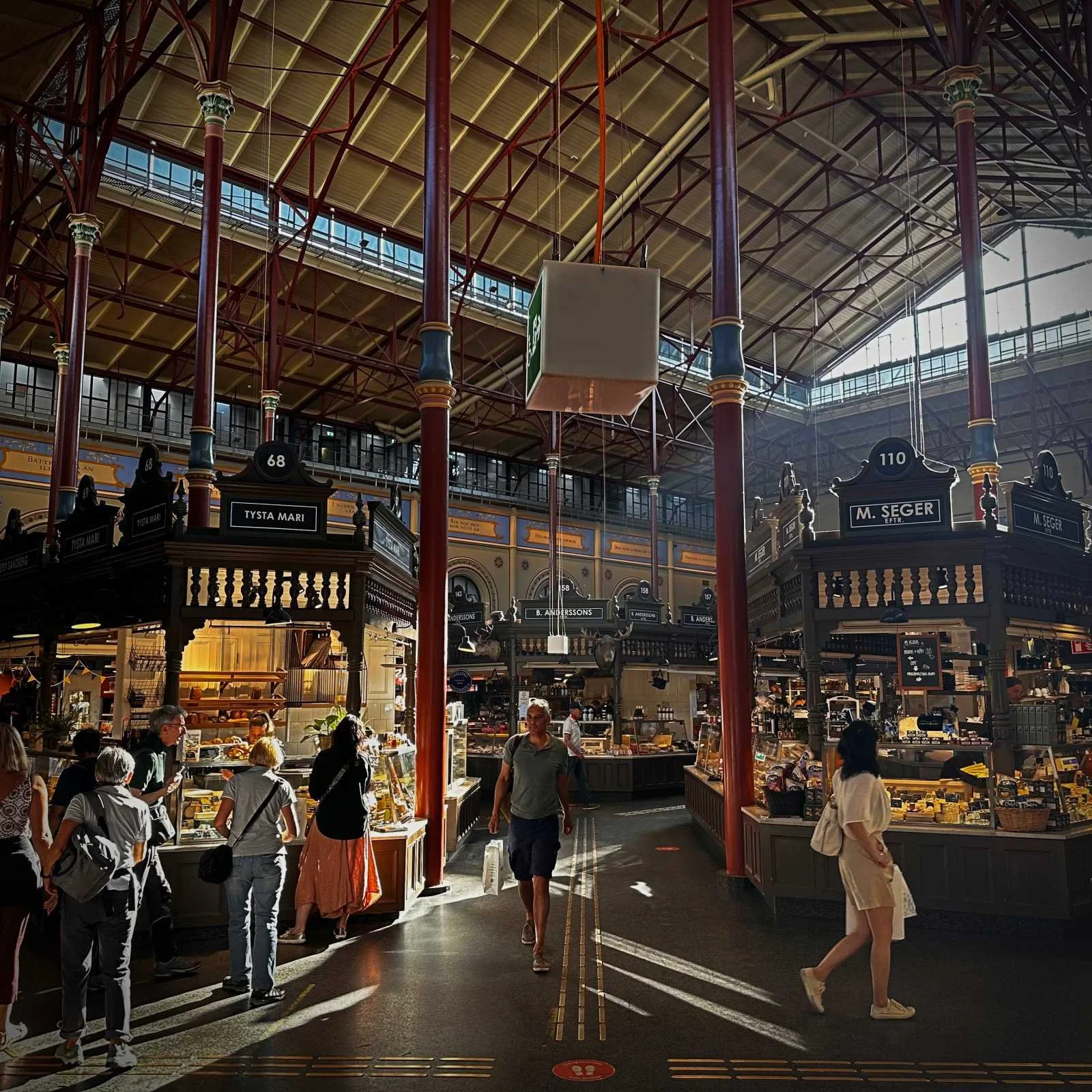 View through the aisles of the historic Östermalms Saluhall in Stockholm. The photo shows the striking red metal ceiling structure, with sunlight falling on wooden market stalls such as "Tysta Mari" and "M. Seger".
