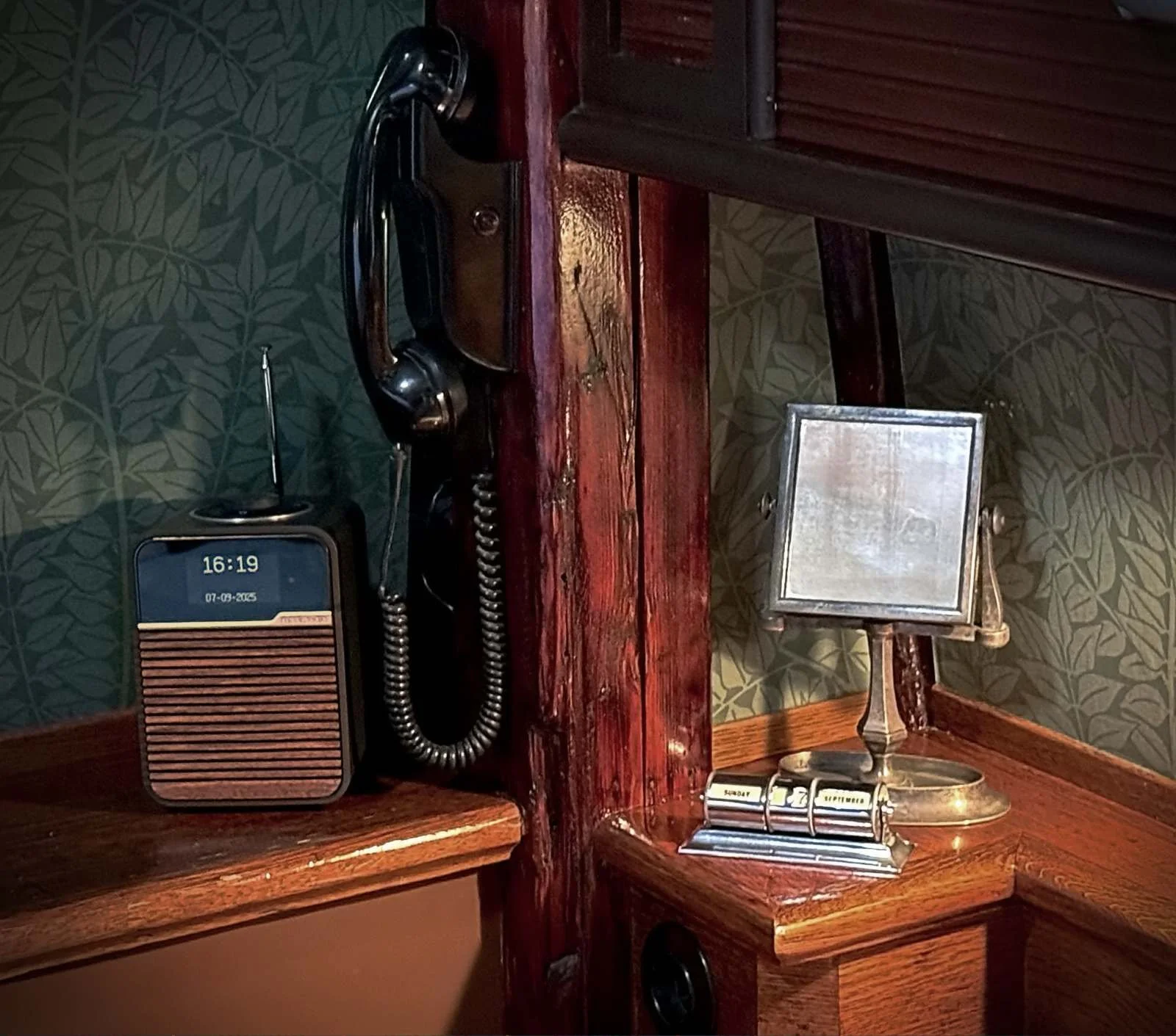 Ein schwarzes Retro-Wandtelefon, ein Digitalradio im Holzdesign, ein kleiner Tischkalender und ein Tischspiegel stehen auf hölzernen Ablagen vor einer grünen Mustertapete.