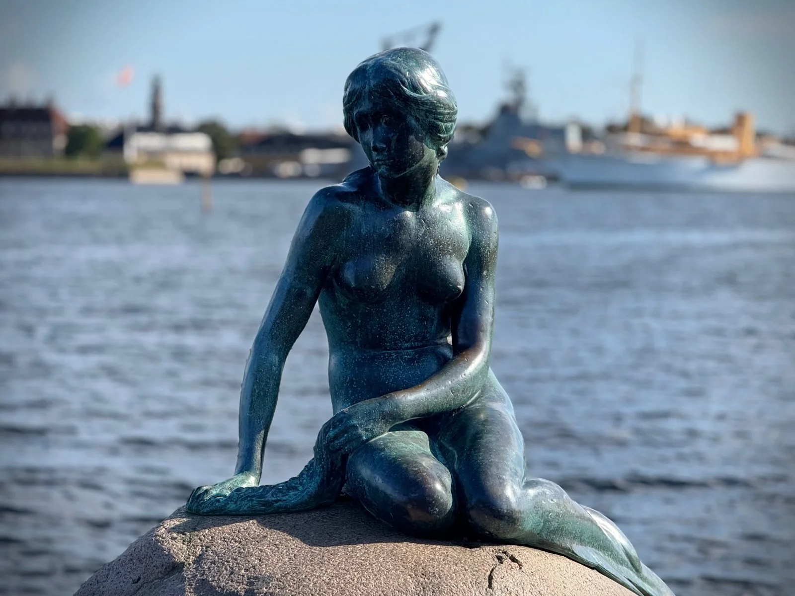 The bronze statue of the Little Mermaid sits on a rock by the shore in Copenhagen. Water is visible in the background.