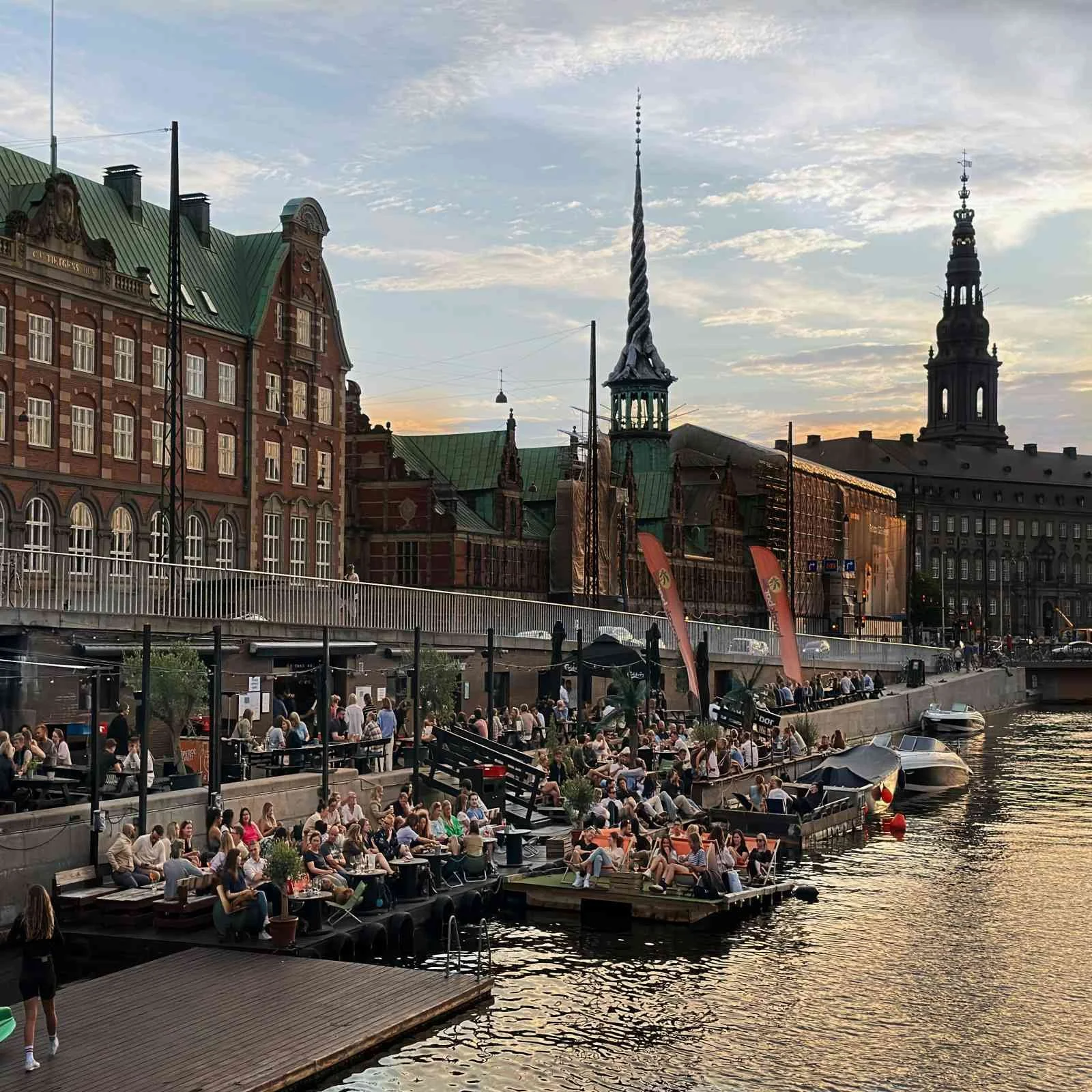 Abendliche Szene an der Kayak Bar in Kopenhagen, Indre By: dicht besetzte Holzdecks am Kanalufer, Boote am Kai, darüber historische Backsteinfassaden mit grünen Dächern und markanten Turmspitzen.