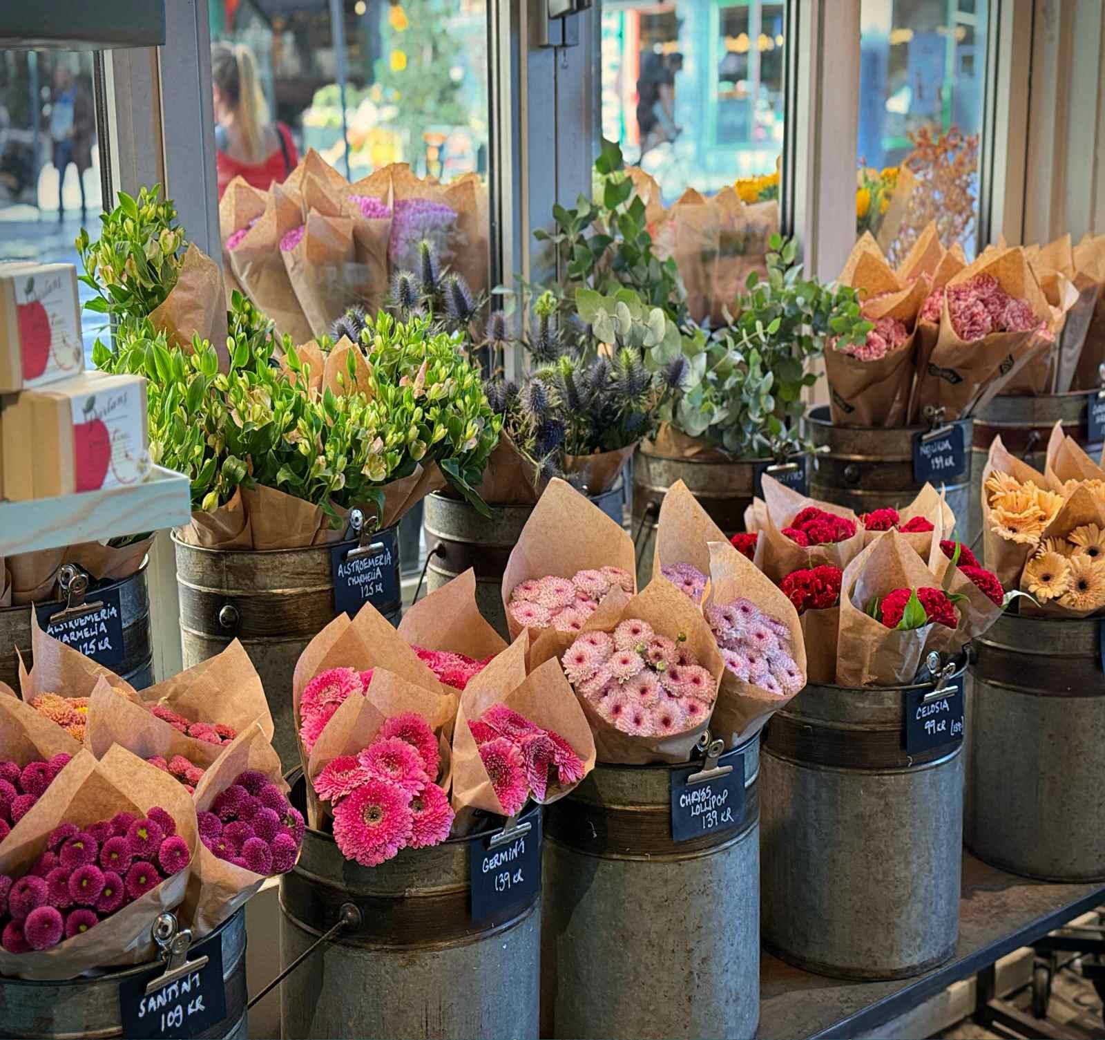 Various flower bouquets wrapped in brown paper stand in zinc metal buckets. Small black clip-on tags on the buckets display the flower names and prices in Swedish Kronor.