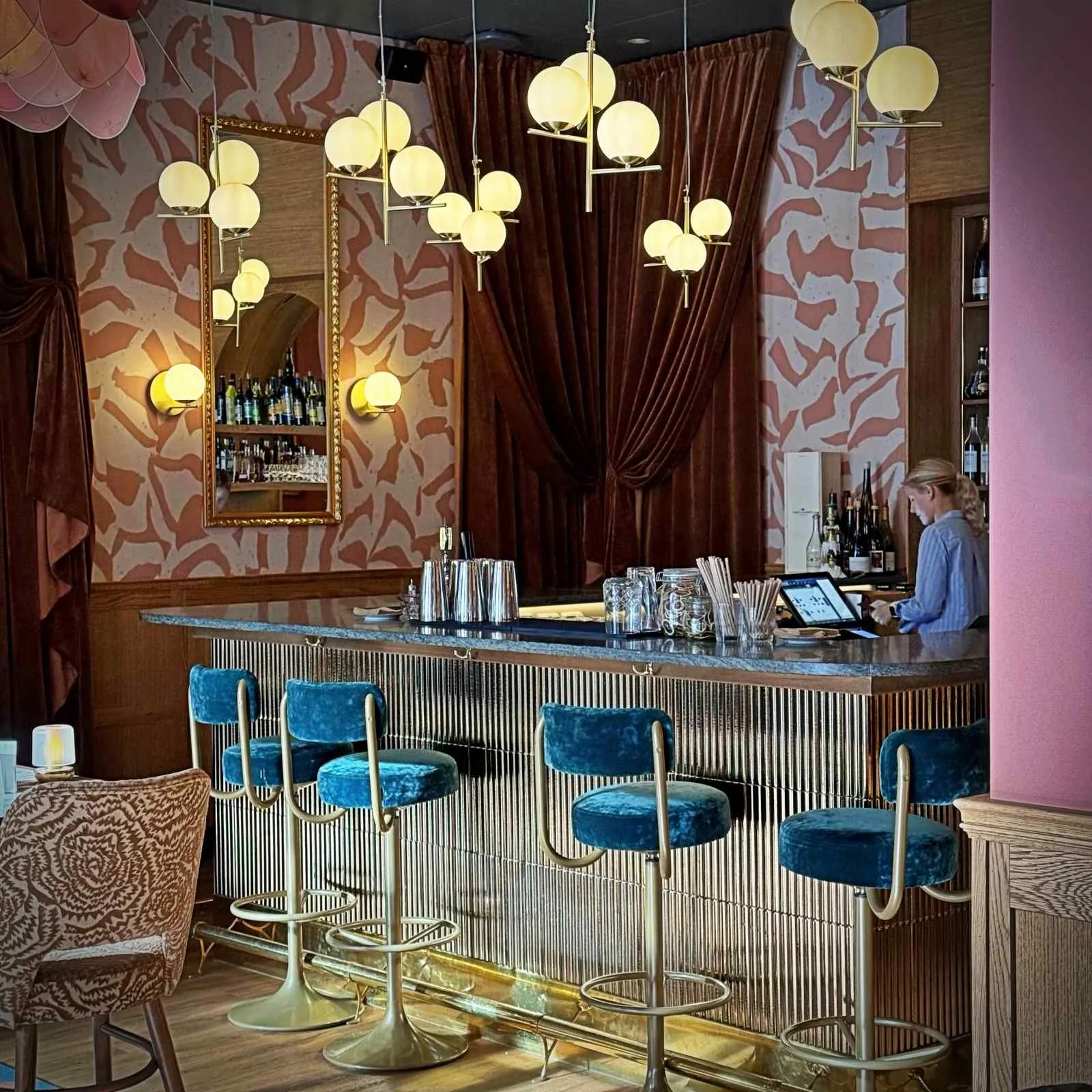 Eine Bar mit einem goldfarbenen Metalltresen, grauer Steinplatte und blauen Samt-Barhockern. Eine Person im gestreiften Hemd steht hinter der Bar an einer Kasse.