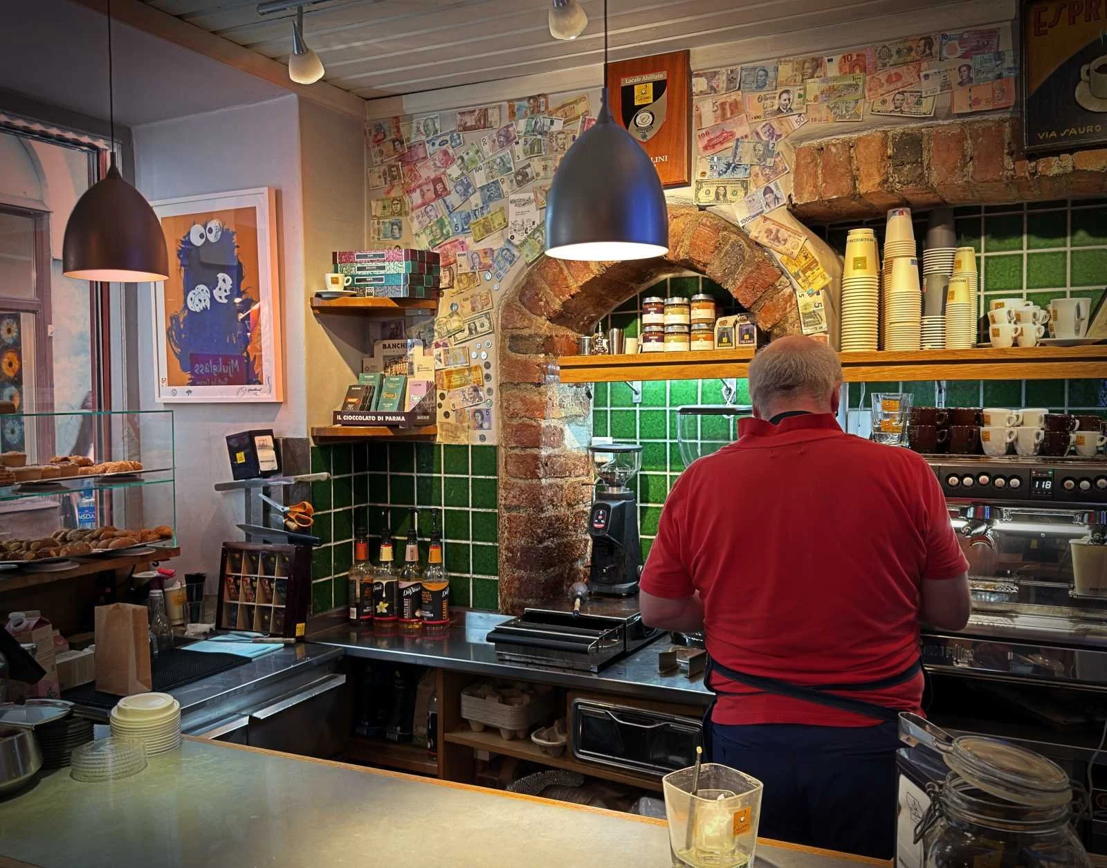 Blick auf den Rücken eines Baristas im roten Shirt an der Espressomaschine vor einer Wand mit Geldscheinen und Backsteinbogen.