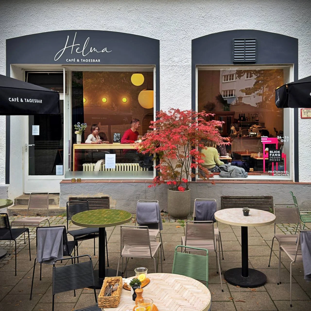 Außenansicht des Café Helma in München mit beschrifteter Fensterfassade, Terrasse mit bunten Stühlen und Tischen, japanischem Ahornbaum und Gästen im Innenraum bei Tageslicht