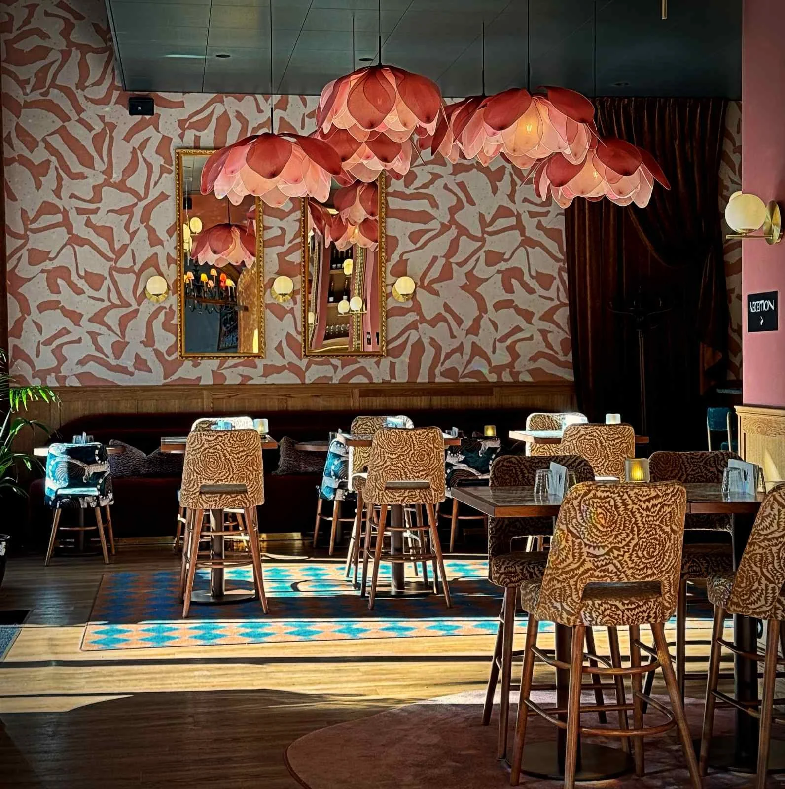 Ein Sitzbereich im The Sparrow Hotel mit Holztischen und gemusterten Barhockern. Von der Decke hängen mehrere rosafarbene Leuchten in Blütenform vor einer gemusterten Wand.