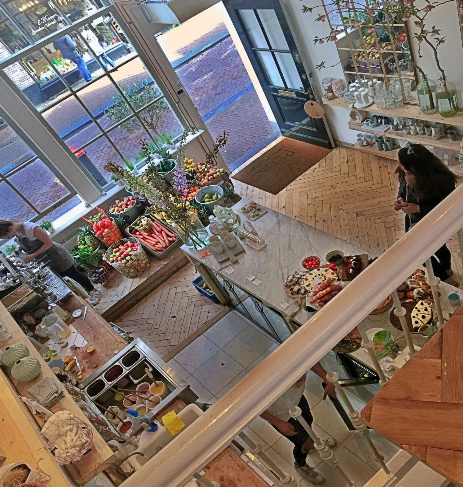 High-angle view inside Café Pluk in the Nine Streets. Baskets of fruit and vegetables stand by the window, while various cakes are arranged on the marble counter.