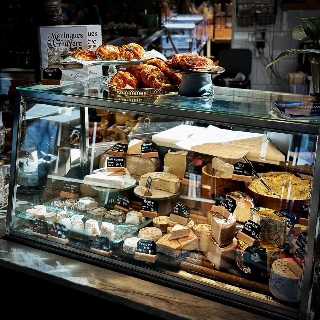 Eine Glasvitrine in der Östermalms Saluhall ist mit verschiedenen Käsesorten wie Saint Agur und Pico bestückt. Auf der Vitrine stehen Etageren mit Zimtschnecken und anderem Gebäck.