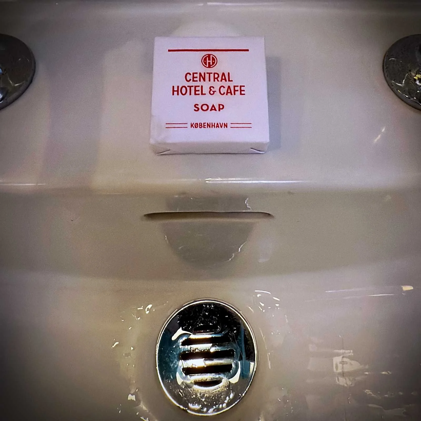 Eine quadratische, weiß verpackte Stückseife mit der roten Aufschrift Central Hotel & Cafe Soap København liegt auf dem Rand eines weißen Waschbeckens über dem silbernen Abfluss.