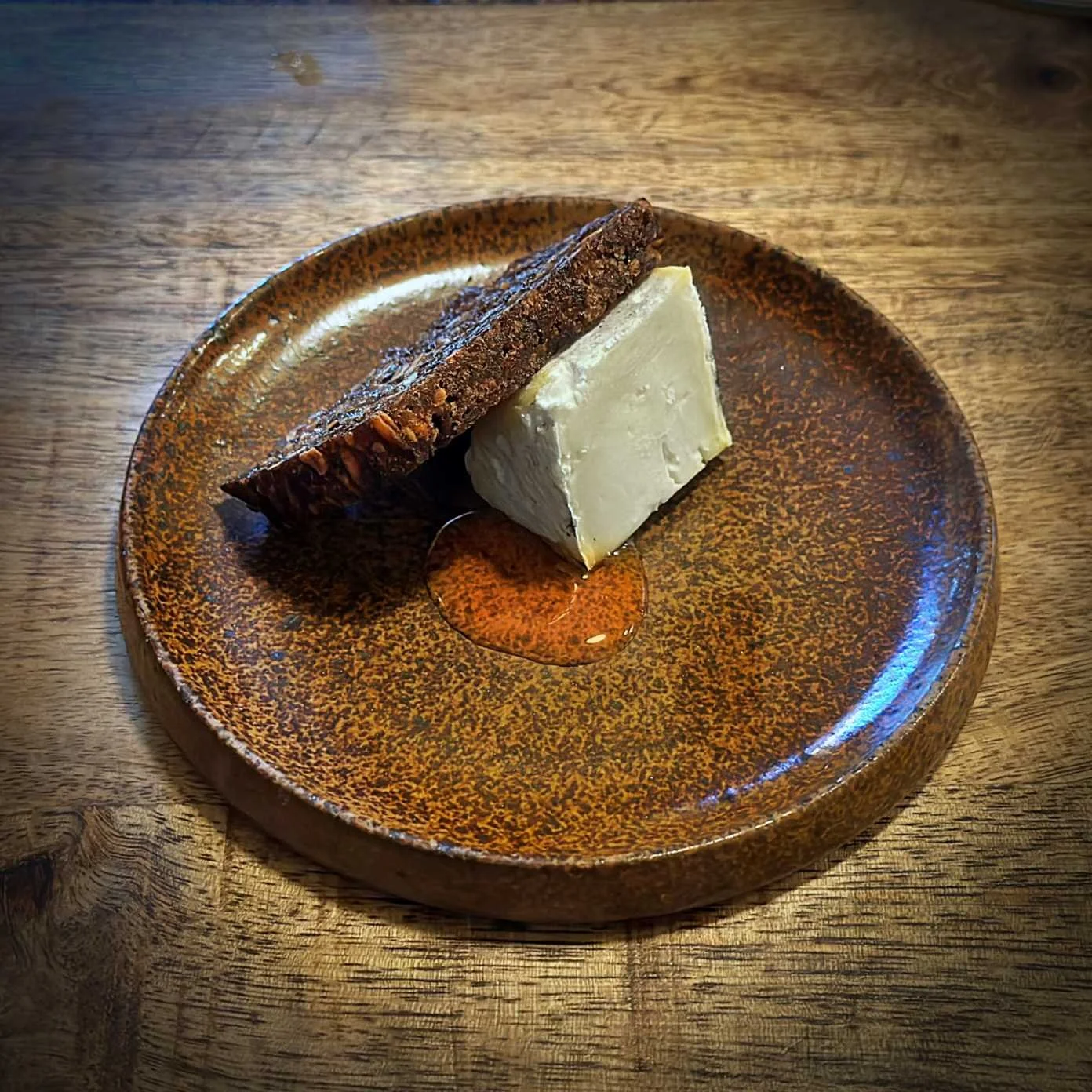 Ein Stück dänischer Aschekäse von Arla Unika mit einer Scheibe dunklem Roggenbrot von der Juno Bakery und Honig auf einem braunen Keramikteller im Restaurant Selma in Kopenhagen. Das Gericht steht auf einem Holztisch.