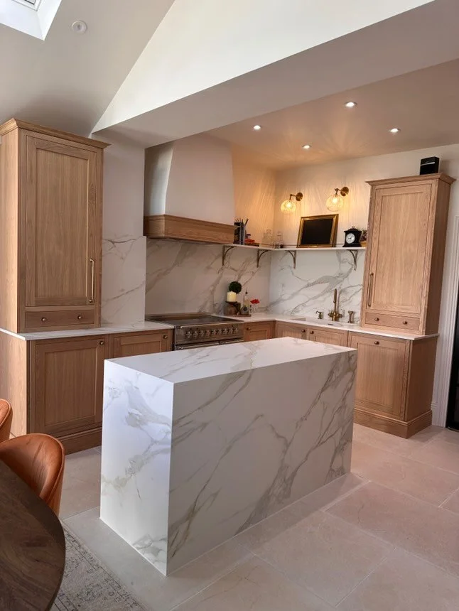 Modern kitchen with wooden cabinets, a marble island, a stainless steel oven, and a marble backsplash. There are two wall-mounted lights and a small chalkboard on the shelf above the sink.