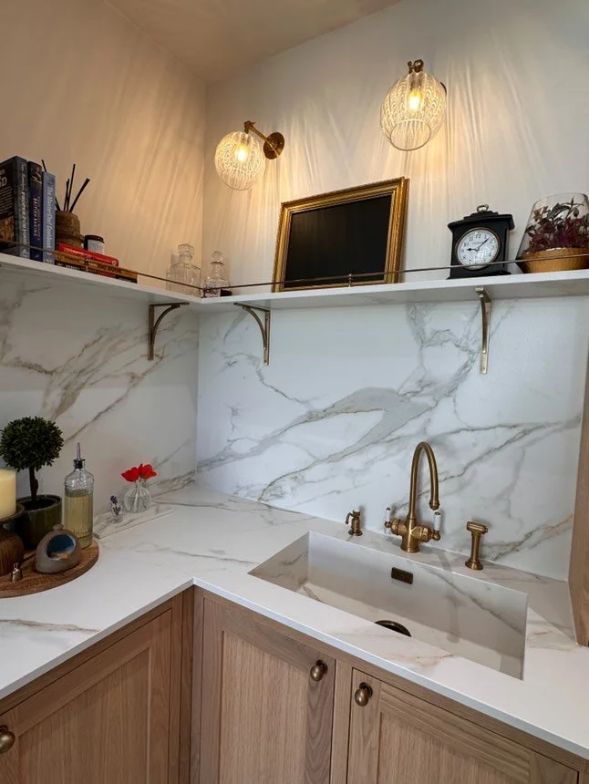 A modern kitchen corner with a marble backsplash and countertop, wooden cabinets, a gold faucet, and decorative items on the counter and shelf, including books, candles, bottles, and a clock.