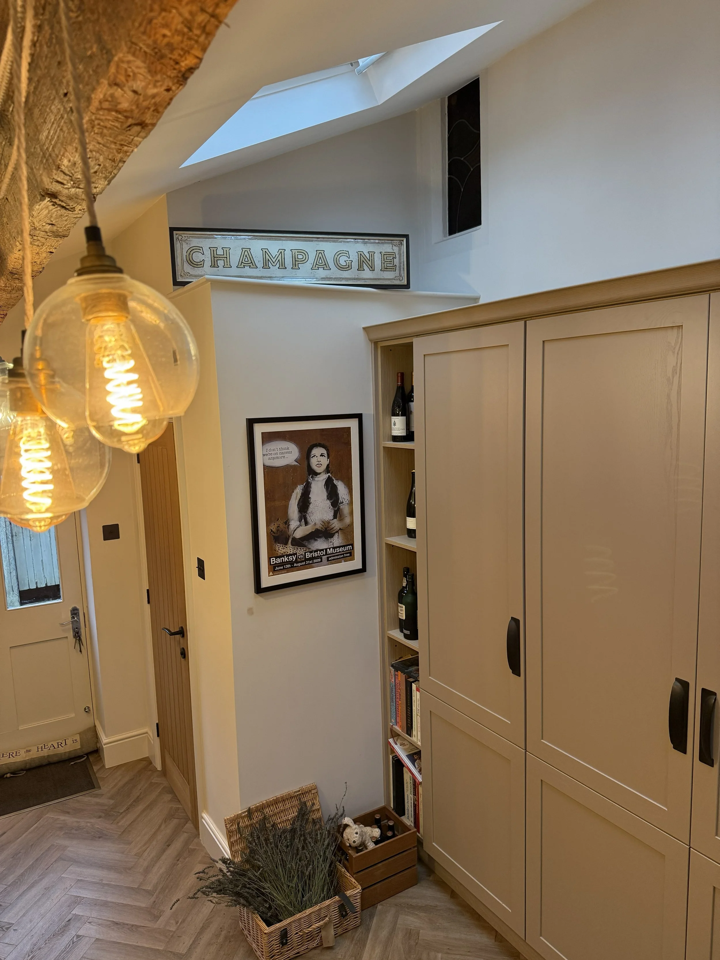 Interior view of a cozy home hallway featuring a large beige storage cabinet, a shelf with wine bottles, framed artwork of a woman with a speech bubble, a wicker basket with plants, and a skylight window on the ceiling. A sign with "CHAMPAGNE" hangs 