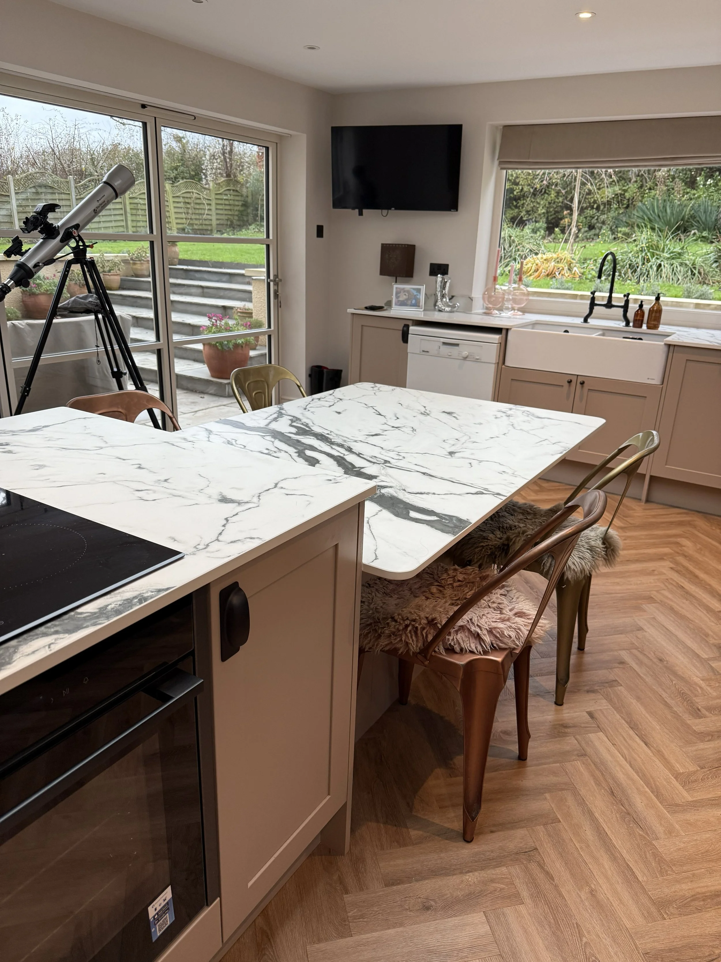 A kitchen with a marble countertop island, a window showing a garden, a telescope pointed outside, a wall-mounted TV, and chairs with fur cushions.
