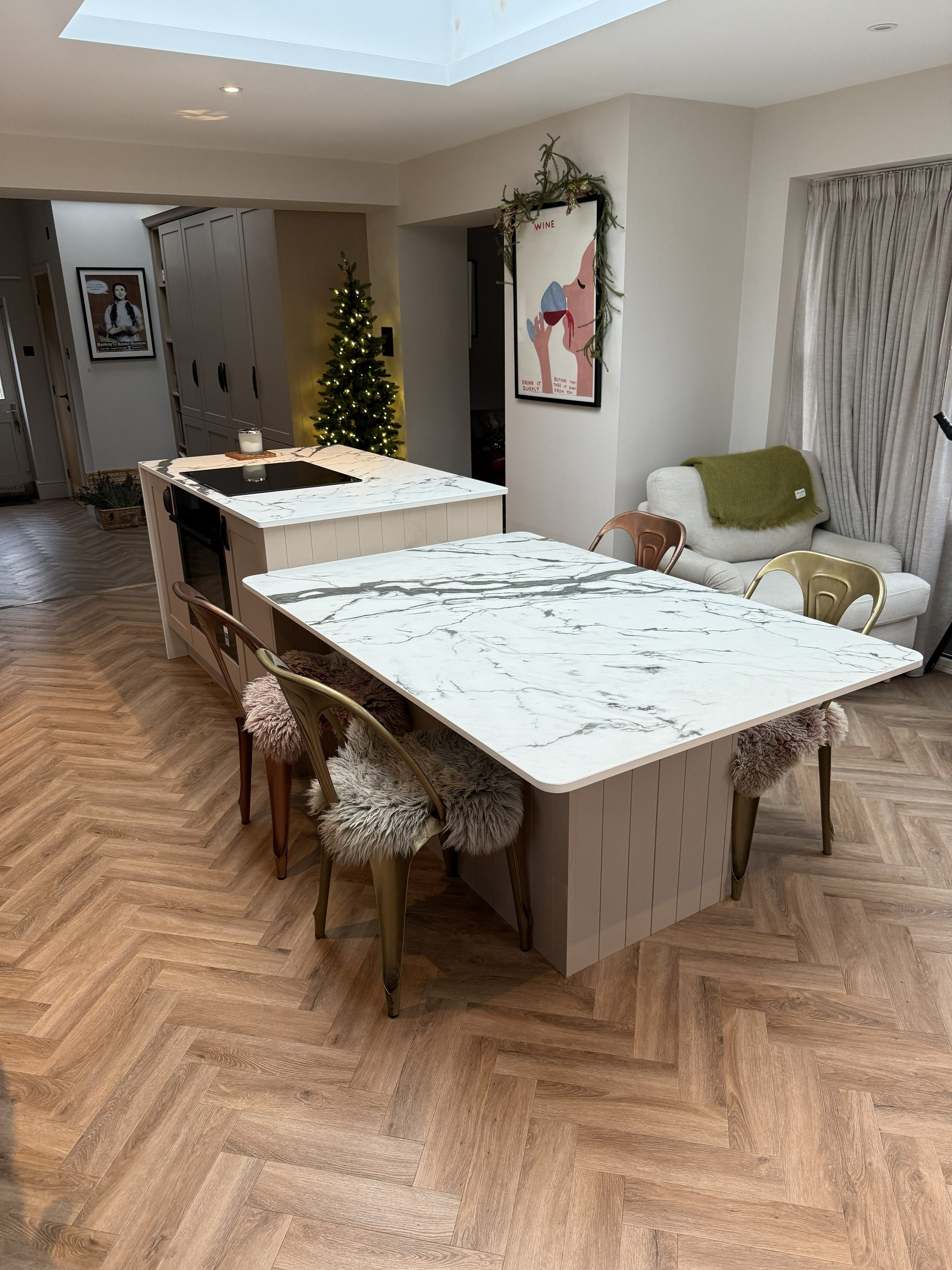 Modern kitchen with island featuring white marble countertop, four chairs with faux fur cushions, and a small Christmas tree in the background. There's artwork of a woman's face on the wall and a window with curtains.