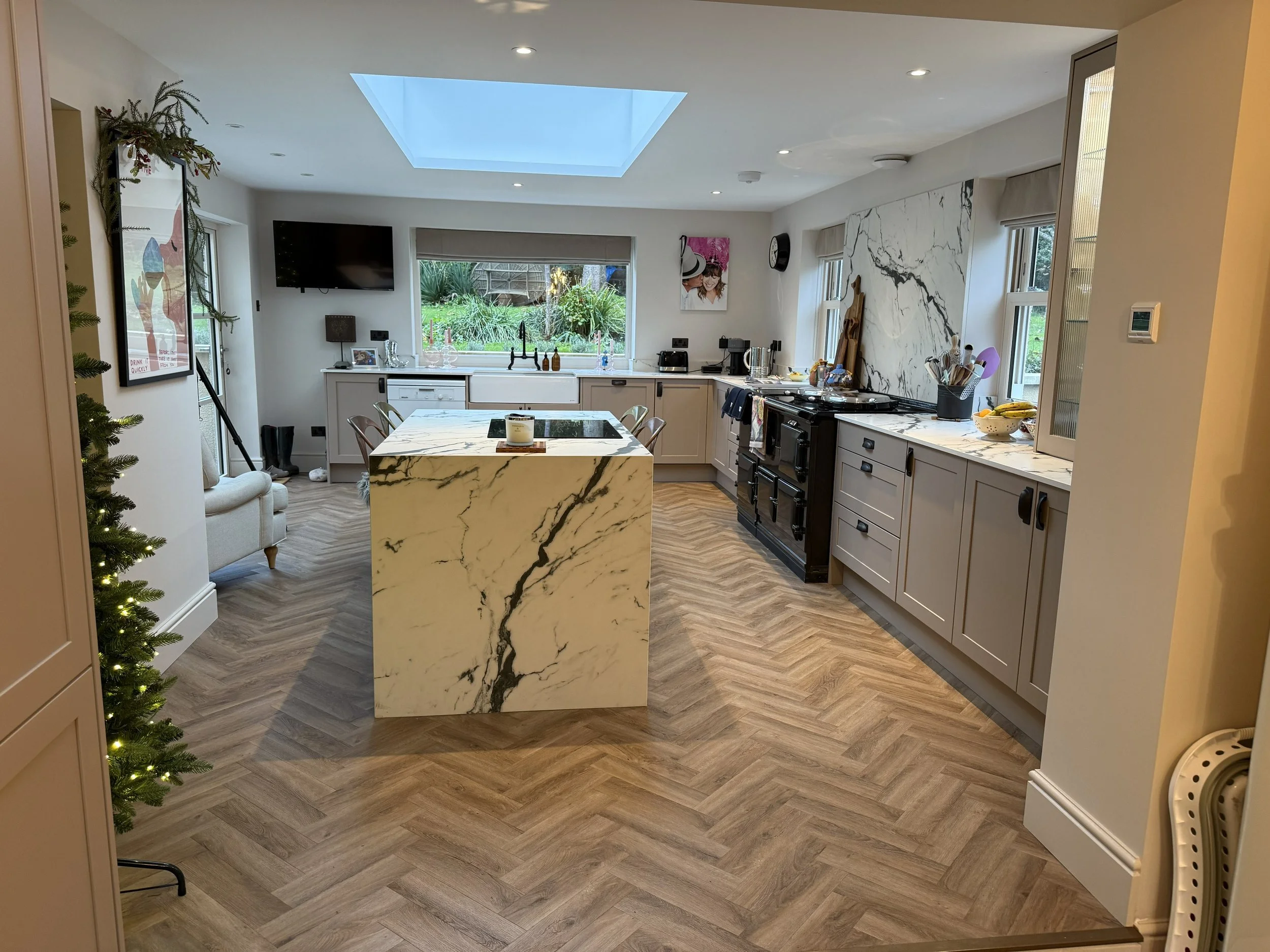 Modern kitchen with marble island, white cabinets, black appliances, and a large skylight above. There's a view of a garden outside the window, a wall-mounted TV, and artwork on the walls.
