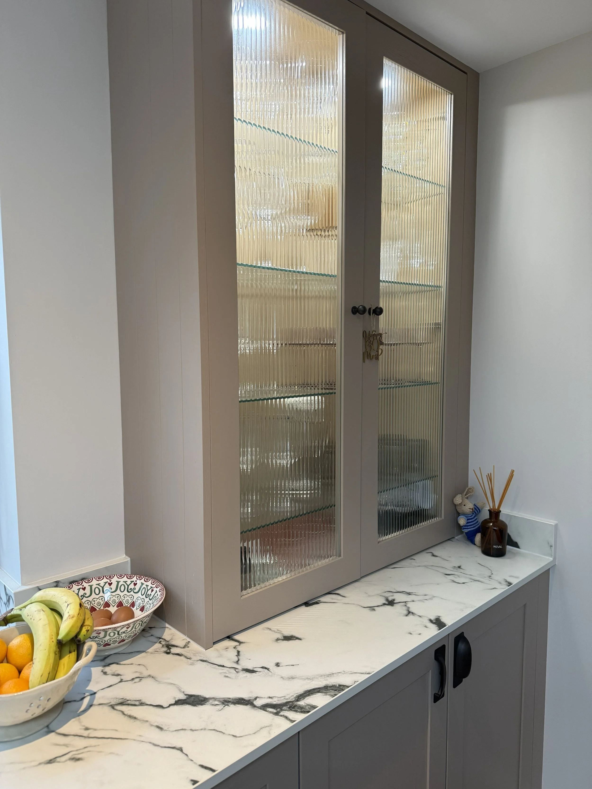 A kitchen countertop with a marble pattern, topped by a bowl of bananas, oranges, and eggs, and a decorative bowl with heart motifs. Above it, a gray cabinet with glass doors containing shelves, and small decorative items including a small stuffed an