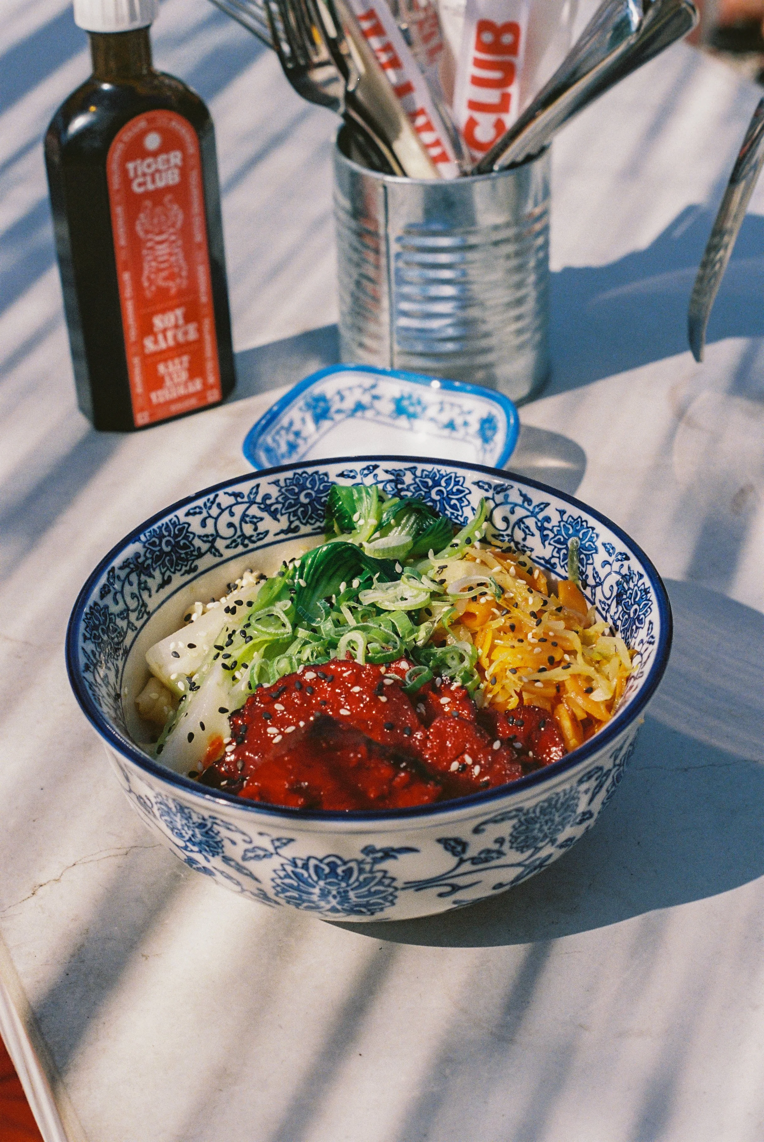 Bol de nourriture asiatique avec pâtes, légumes verts, sauce rouge, et graines de sésame, placé sur une table avec condiments et ustensiles.