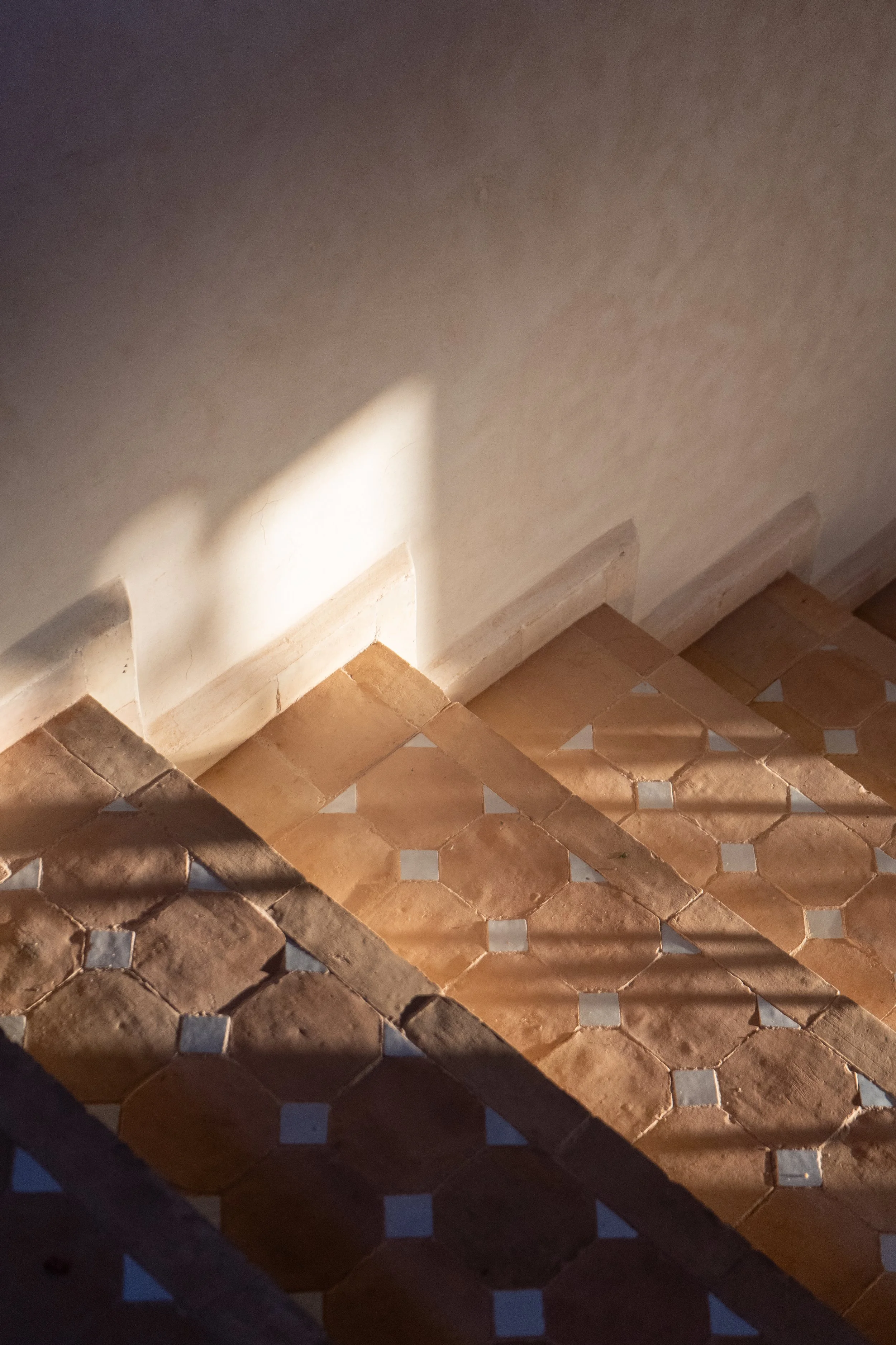 Lumière naturelle projetée sur un mur blanc et un sol en carreaux de terre cuite avec motifs blancs, créant des ombres et des jeux de lumière.