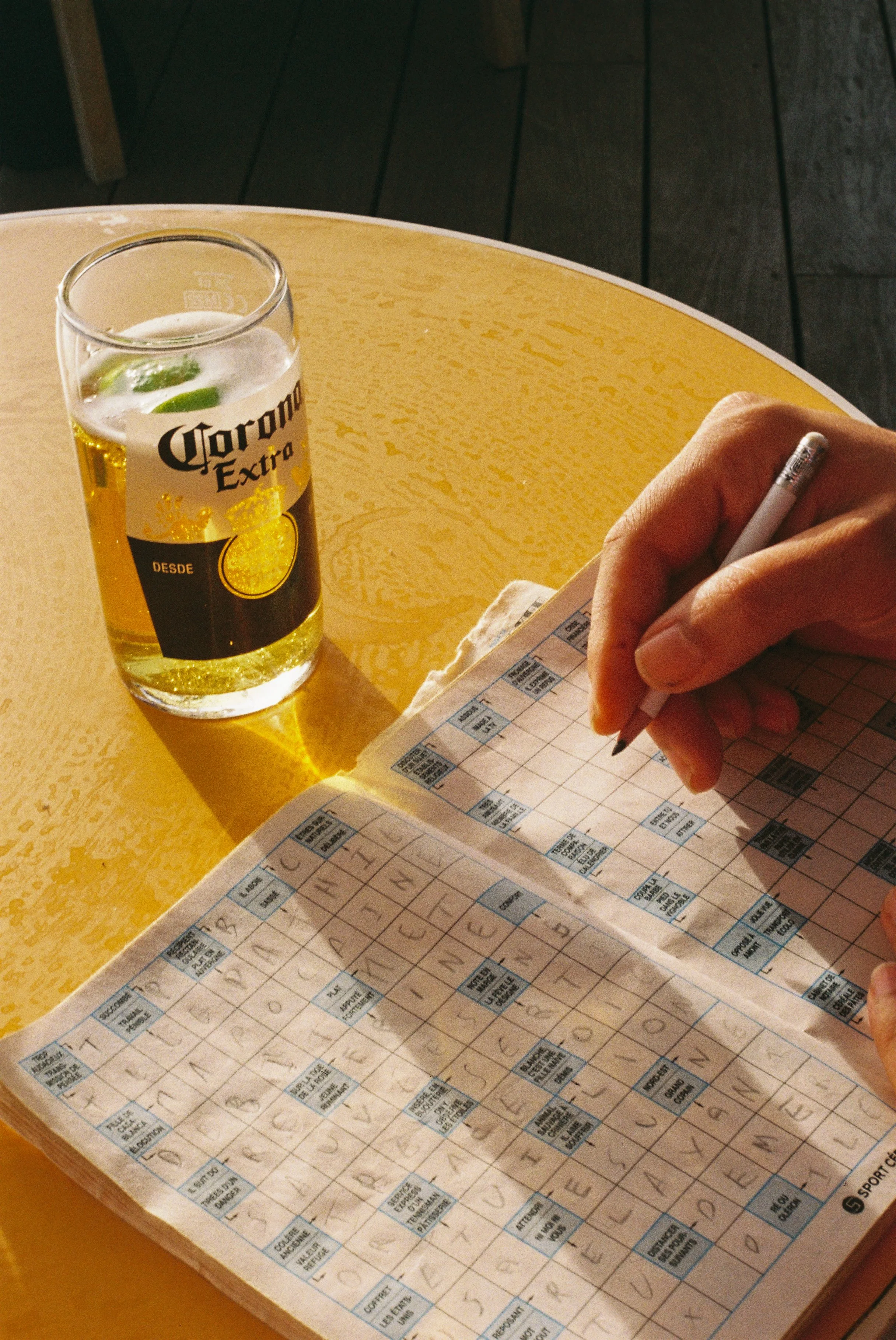 Une personne remplit une grille de mots croisés sur une table jaune, avec une bière Corona à côté dans un verre.