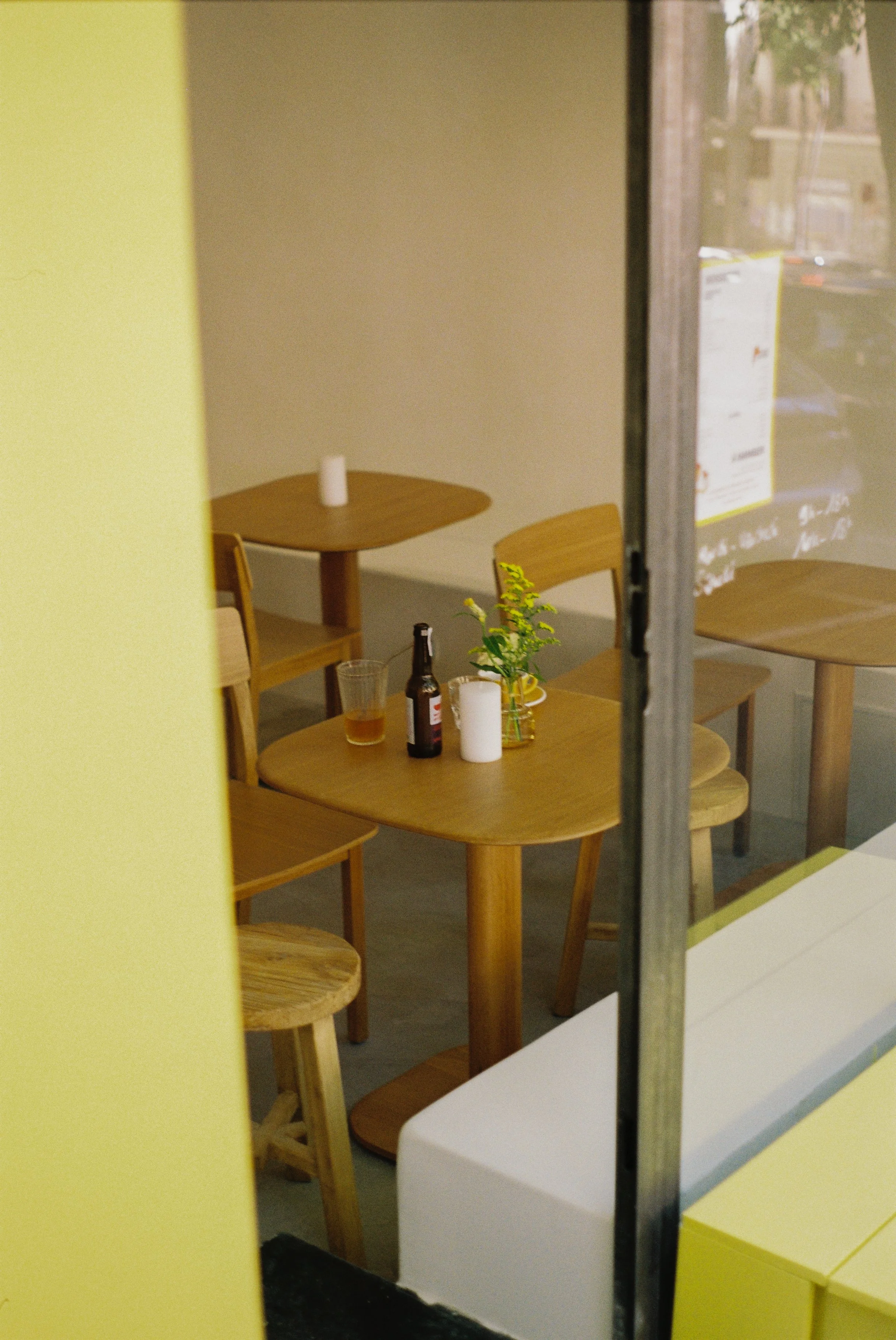 Intérieur d'un café avec une table en bois, une bouteille, un verre, une bougie et un vase avec des fleurs, vue à travers une vitre.