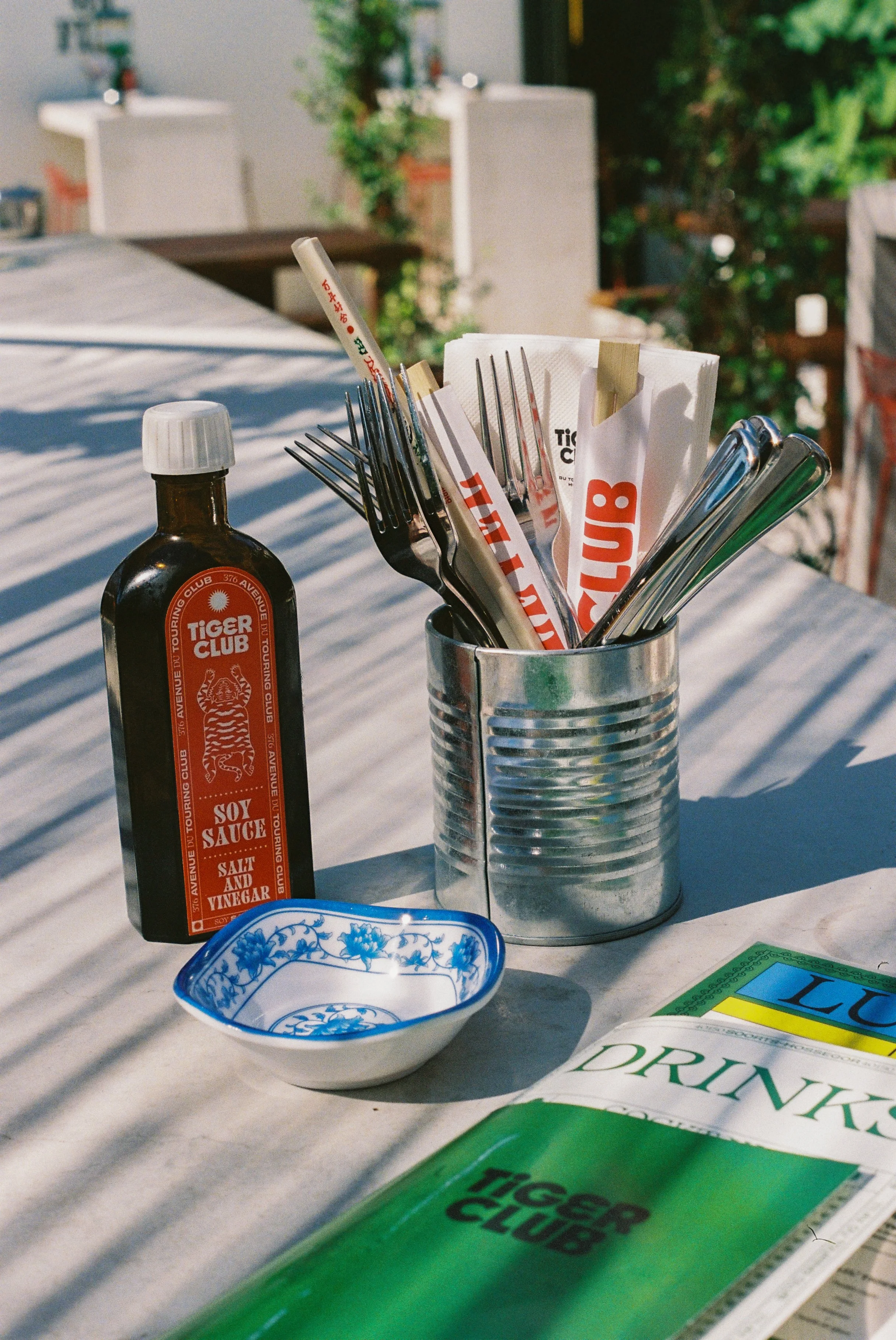 Ustensiles de table dans un porte-couverts en métal, bouteille de sauce soja, bol en porcelaine blanche et bleue, menu avec le logo du Tiger Club, en extérieur avec une table en toile de fond
