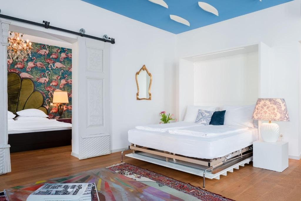 A bedroom with white walls and a blue ceiling with cloud-shaped cutouts. The bed has white bedding and pillows, situated in front of a large wall niche. To the right of the bed, there is a white nightstand with a table lamp featuring a floral lampshade. On the left side, a sliding barn door with a flamingo-themed wallpaper behind it leads to an adjacent bedroom, which has a bed with a tufted, palm leaf-shaped headboard and a bedside table with a lamp. A small gold mirror hangs on the wall between the rooms.