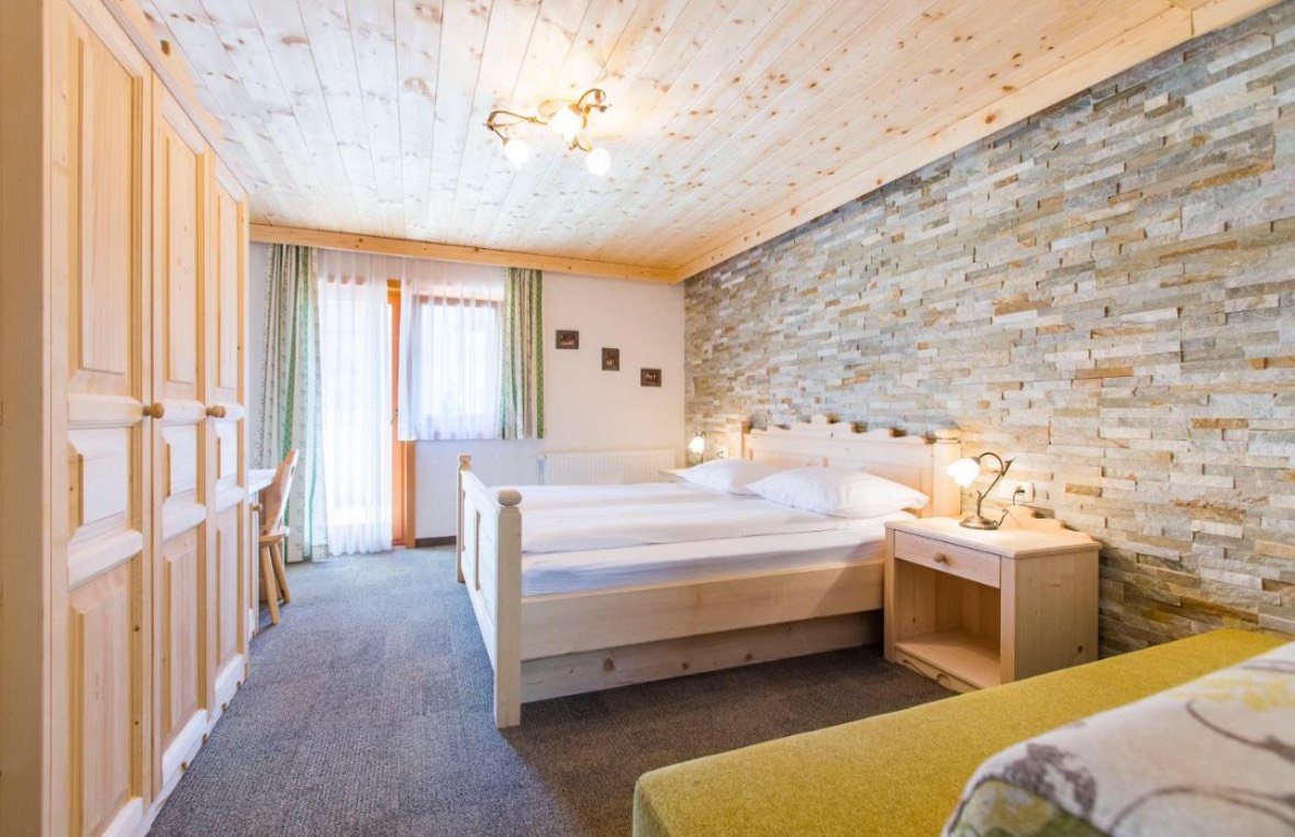 A cozy hotel room with wooden furniture, double beds, a stone accent wall, a wooden wardrobe, a window with curtains, and a lampshade on the nightstand.