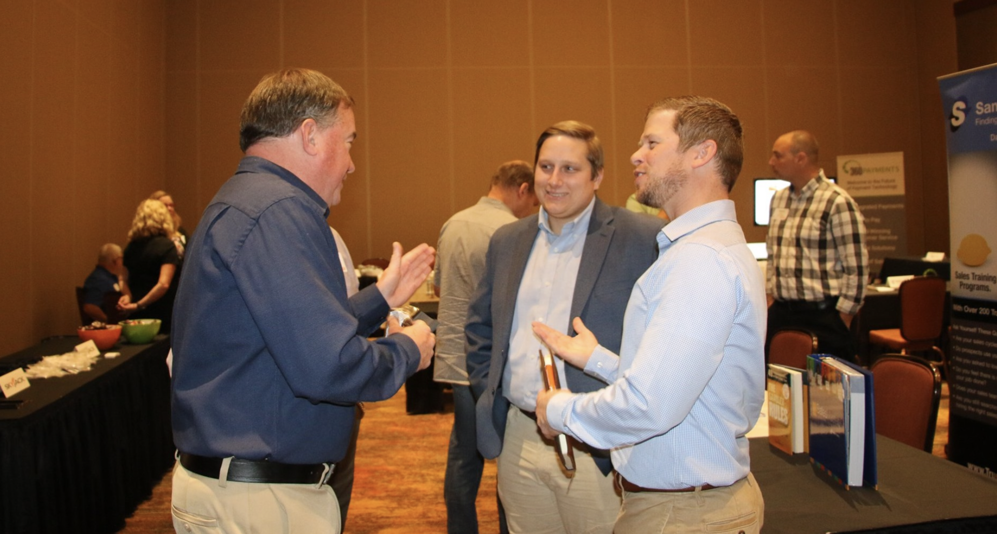 Three men having a conversation at a business event, with an exhibition table and promotional posters in the background.