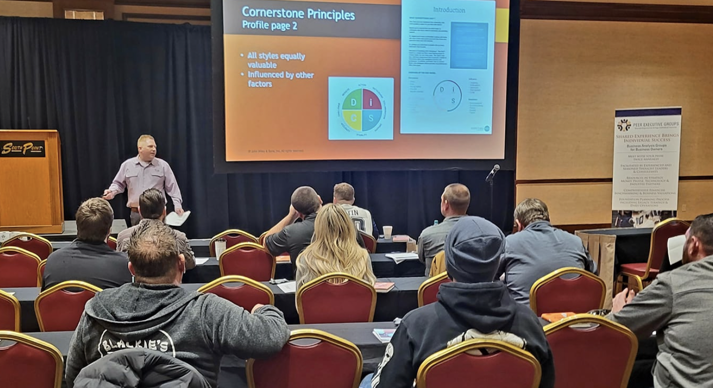 A presentation in a conference room with a man standing in front of a large screen displaying a slide titled 'Cornerstone Principles Profile page 2' with bullet points and diagrams. Audience members seated in red chairs are watching the presentation.