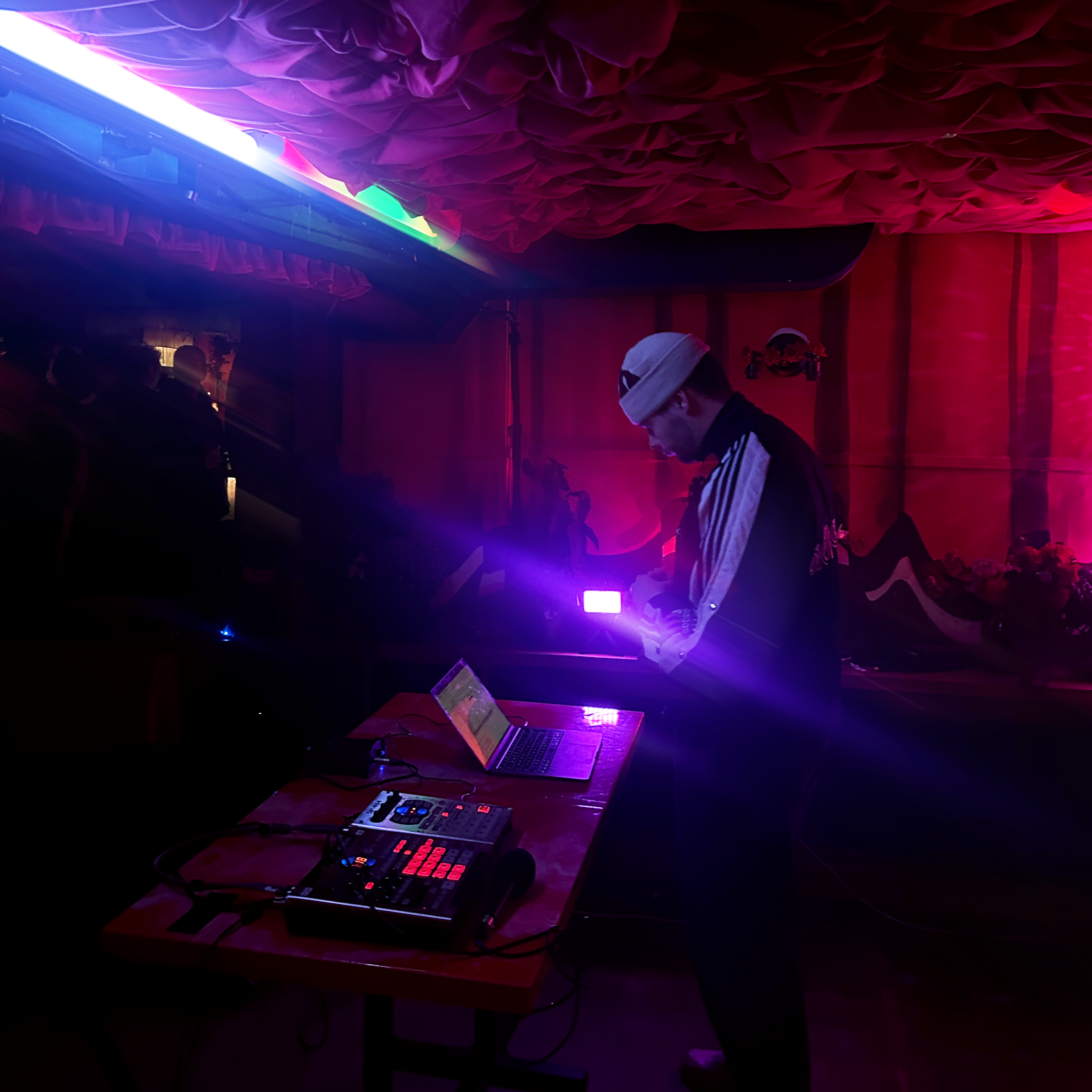 A dimly lit room with purple and pink lighting, showing a person with a white head covering standing near a table with electronic music equipment, including a mixer and a laptop.