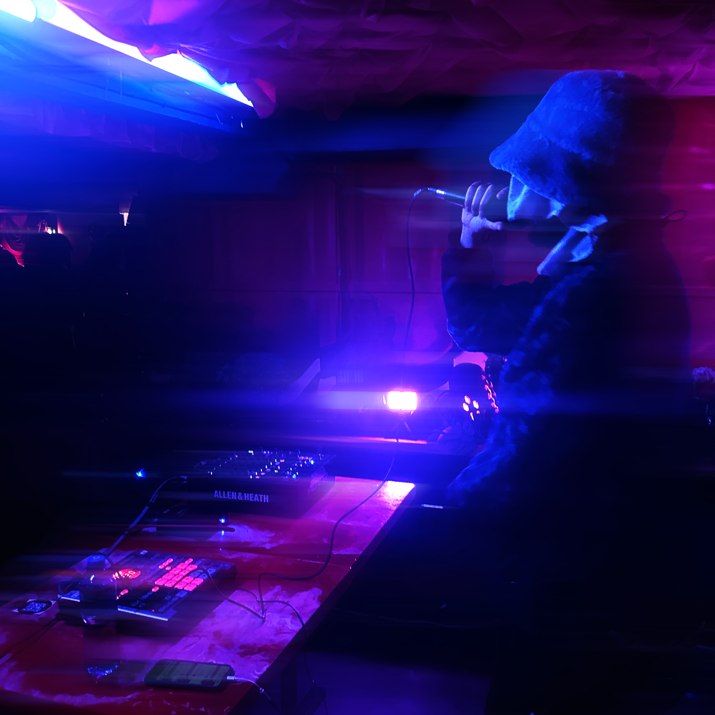 A DJ performing at a nightclub with colorful blue and purple lighting, a mixing console with glowing controls, and on-screen graphics displaying the brand name 'ALLEN & HEATH'.