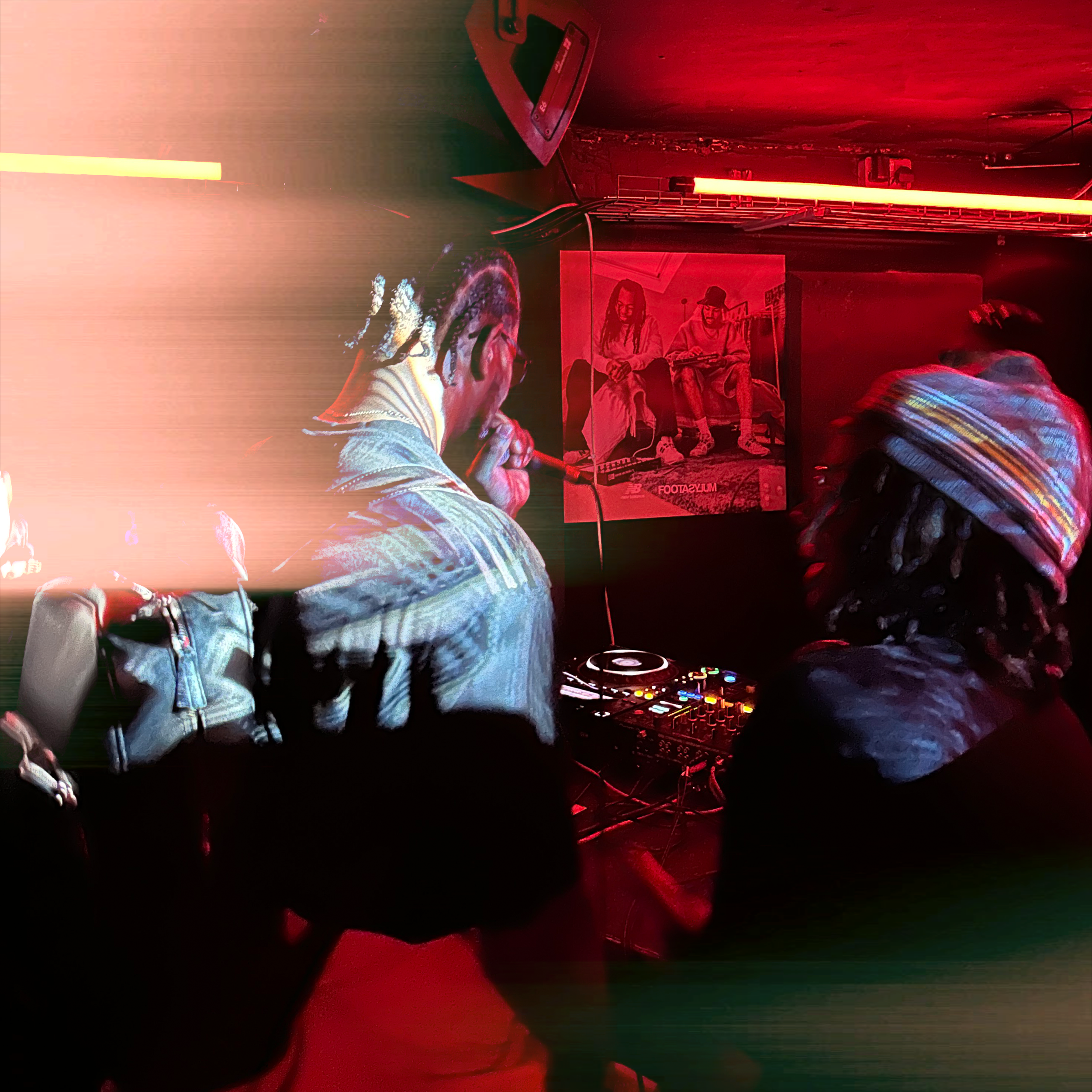 A person with braided hair wearing a jacket, operating DJ equipment in a dimly lit room with red lighting, featuring posters on the wall and a bed.